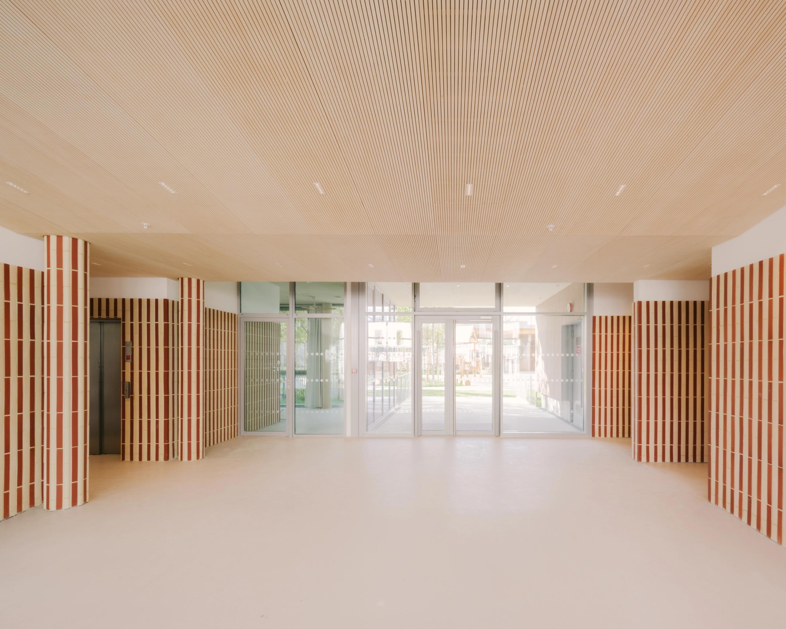 Hall du Groupe scolaire Dominique Frelaut à Colombes, espace traversant lumineux, plafond bois, poteaux en briques verticales et façade vitrée sur cour.