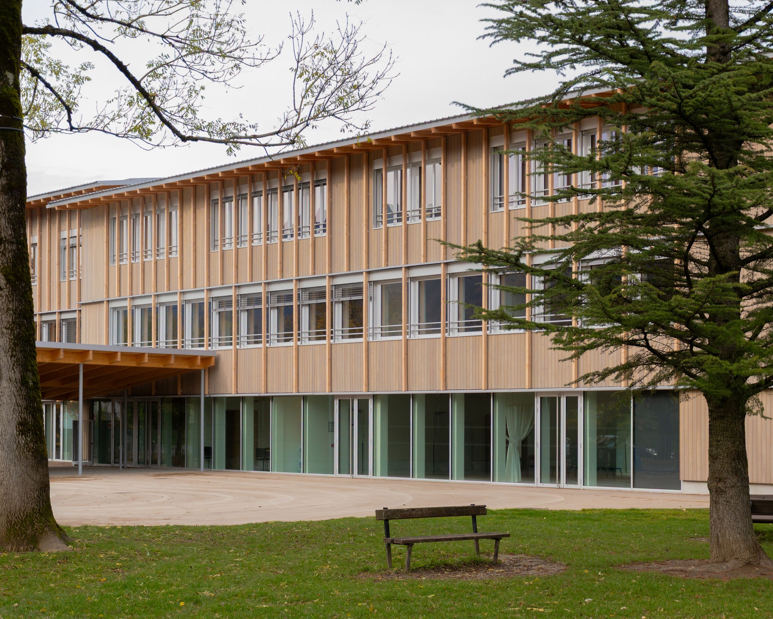 Façade bois surélevée du groupe scolaire du Bouchet à Bonneville, grandes baies vitrées sur cour minérale couverte, en lien direct avec le parc arboré.