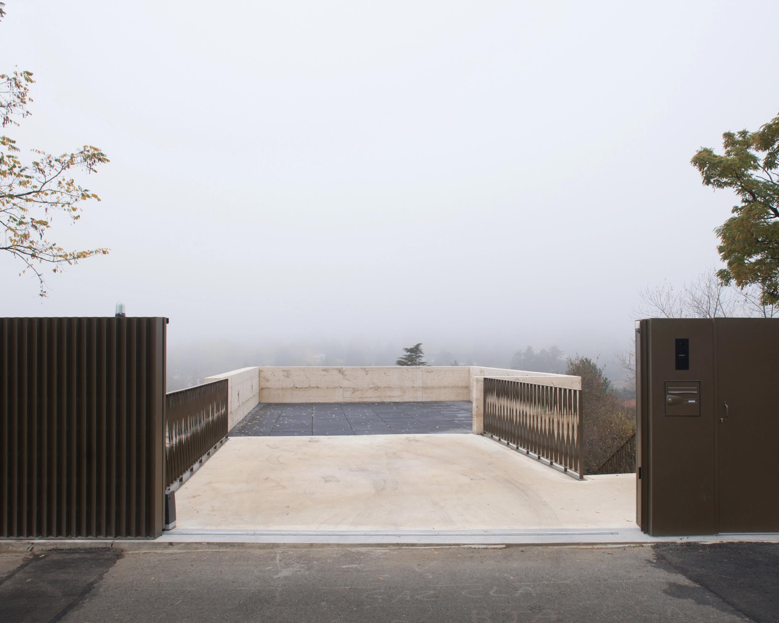 Terrasse d’accès de la maison à Saint-Cyr-au-Mont-d’Or, plateforme en béton teinté encastrée dans le vallon, garde-corps métalliques cadrant le paysage brumeux.