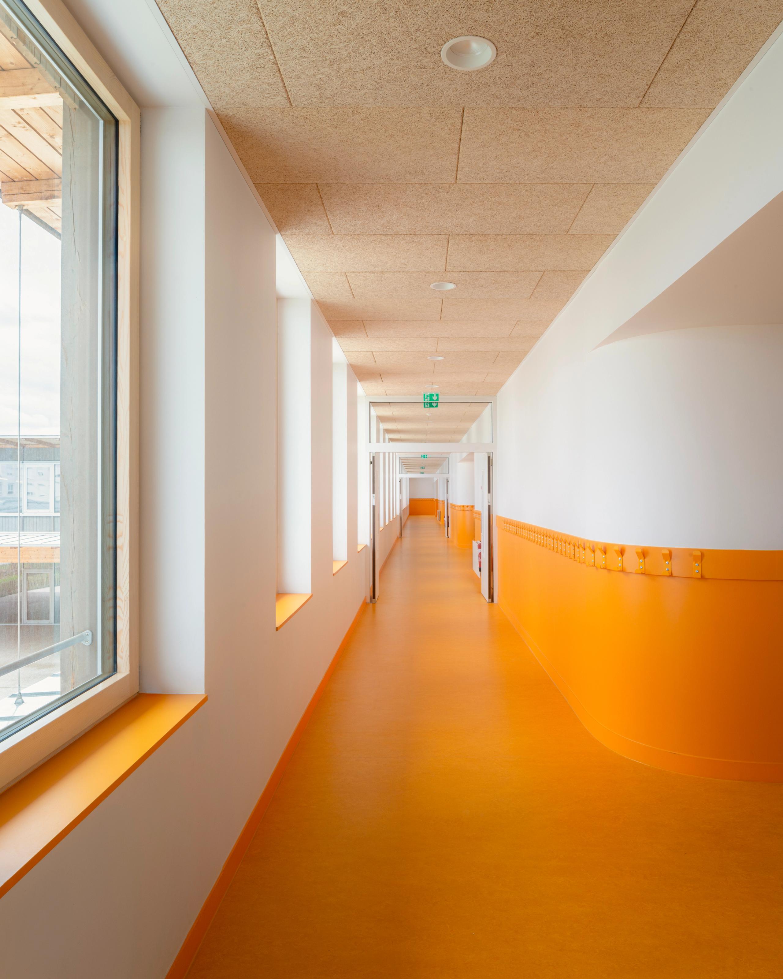 Couloir des groupes scolaires Jacqueline Auriol et Jean Jaurès au Bourget, sol et soubassement orange, plafond bois, grandes baies vitrées sur le parc.