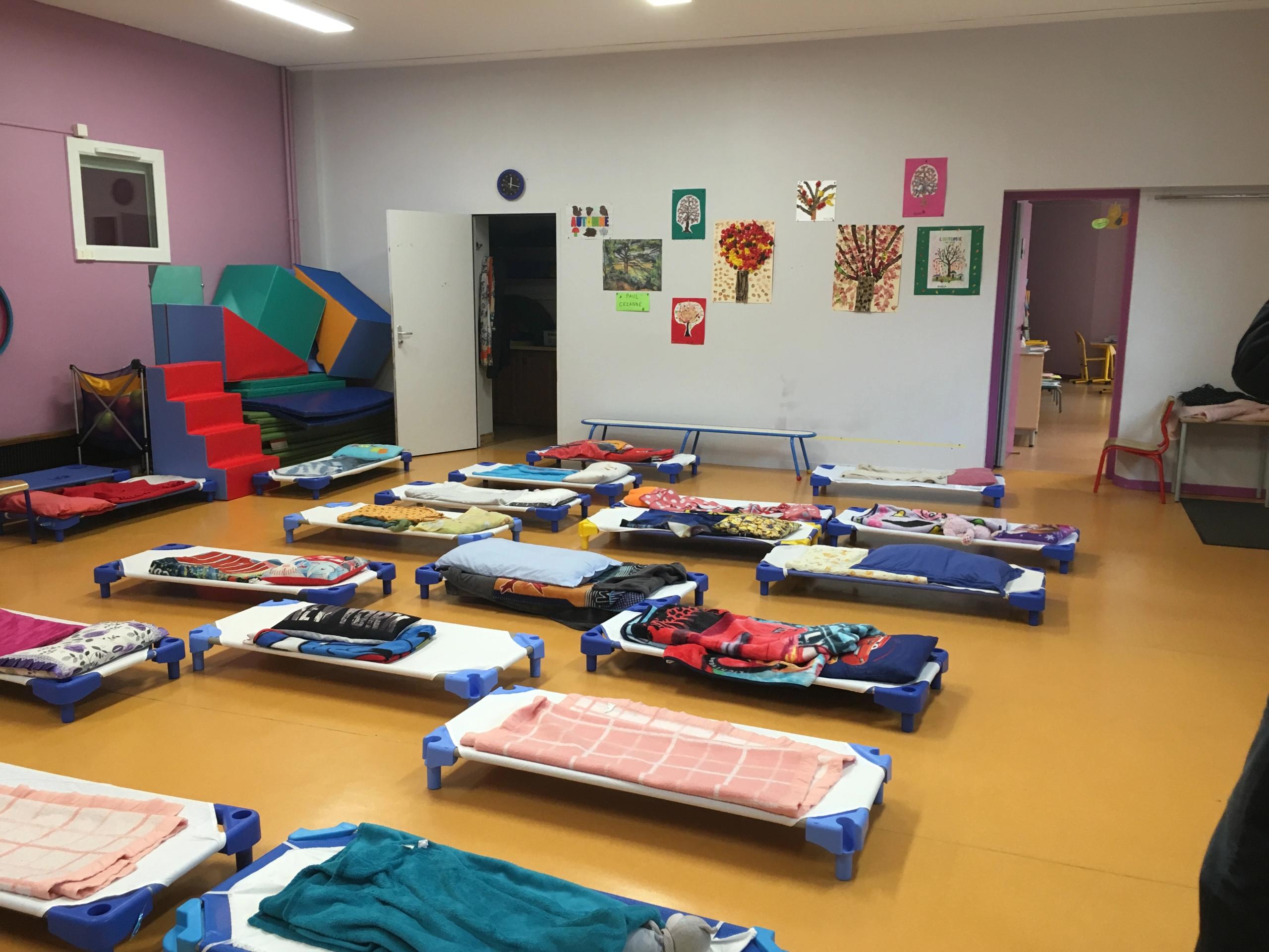 Salle de repos existante du groupe scolaire du Bouchet à Bonneville, avec lits bas alignés sur sol souple, parois colorées et ouverture vers les espaces pédagogiques.