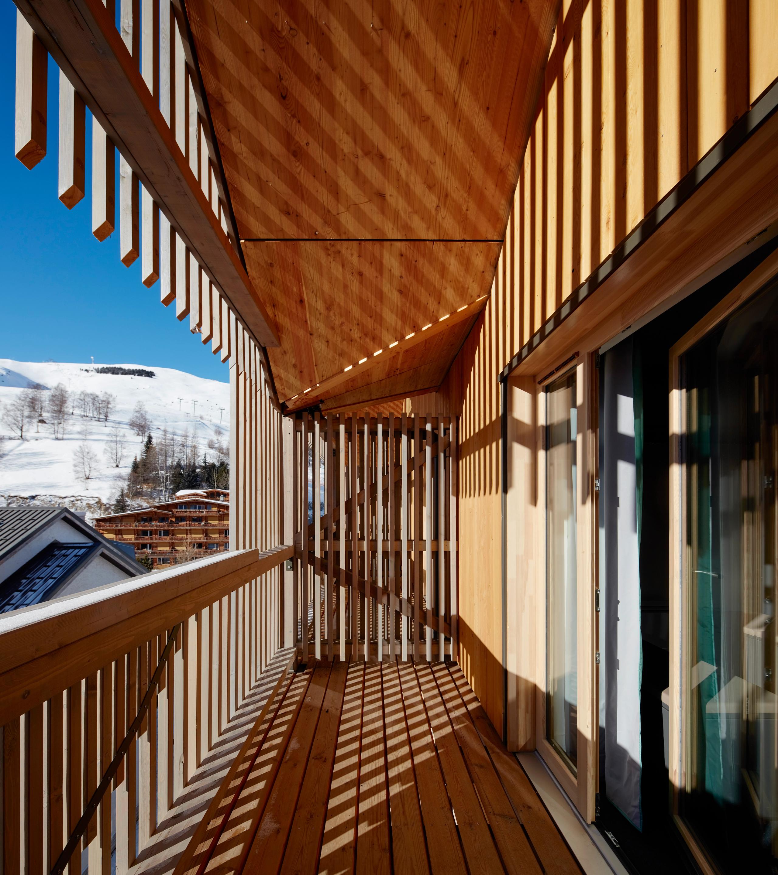 Galerie en bois à claire-voie de l’auberge de jeunesse des Deux Alpes, double peau filtrant la lumière et cadrant le paysage enneigé en arrière-plan.