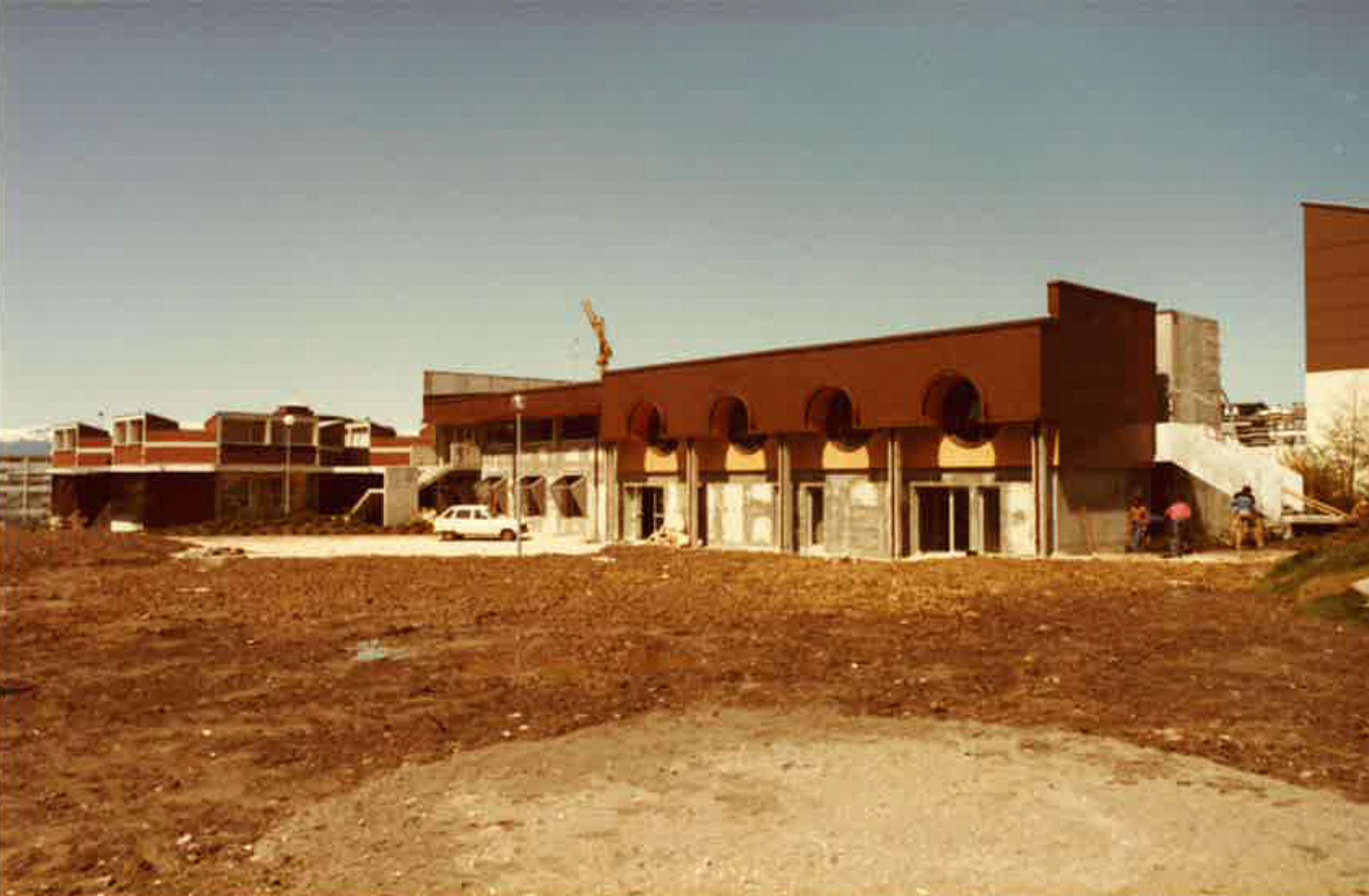 Chantier du conservatoire intercommunal d’Annemasse dans les années 80 : façade en béton, parement brun, baies cintrées et volumes étagés autour d’un terrain nu.