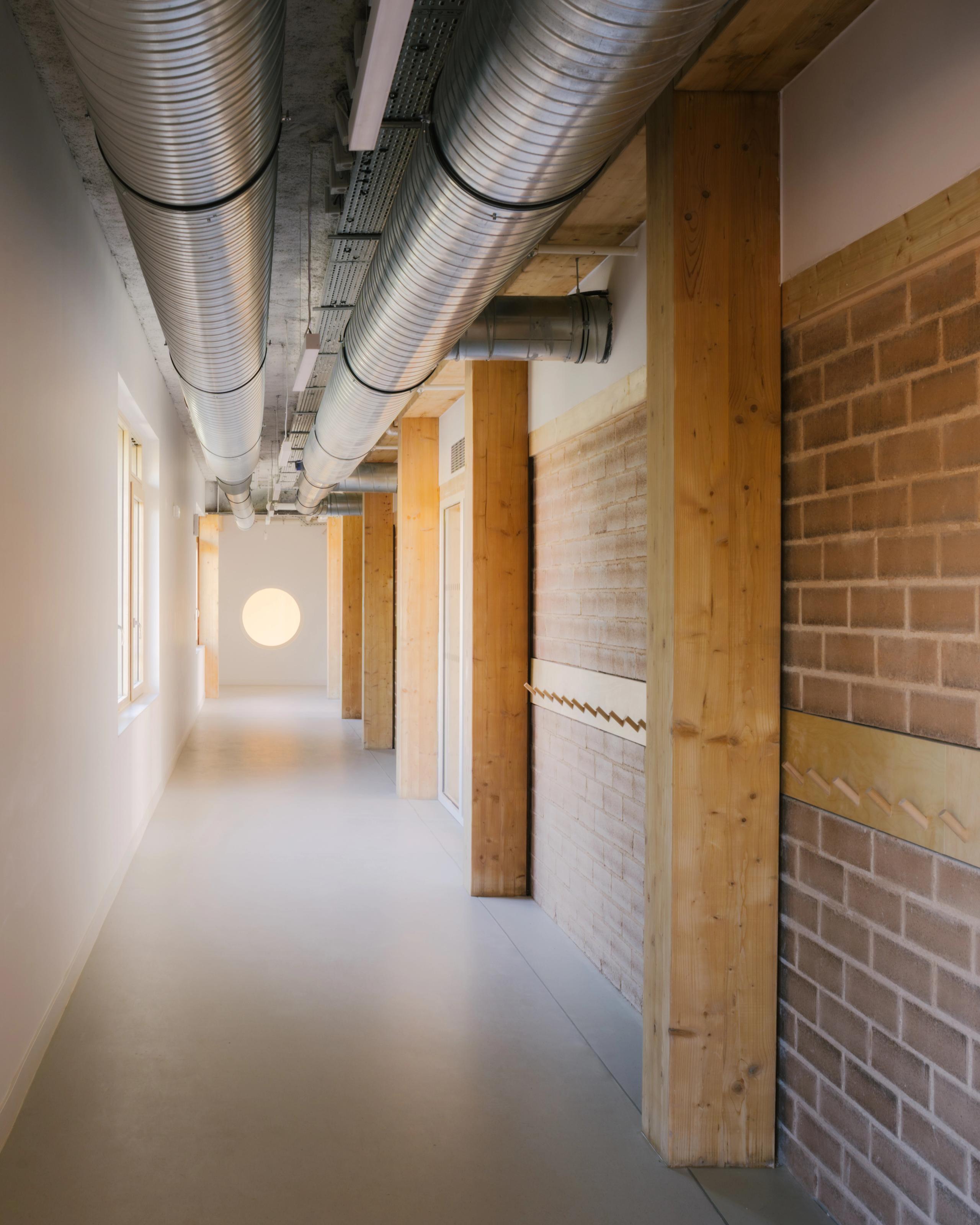 Couloir du groupe scolaire Dominique Frelaut à Colombes, structure bois-brique apparente, gaines techniques visibles et lumière naturelle en bout de perspective.