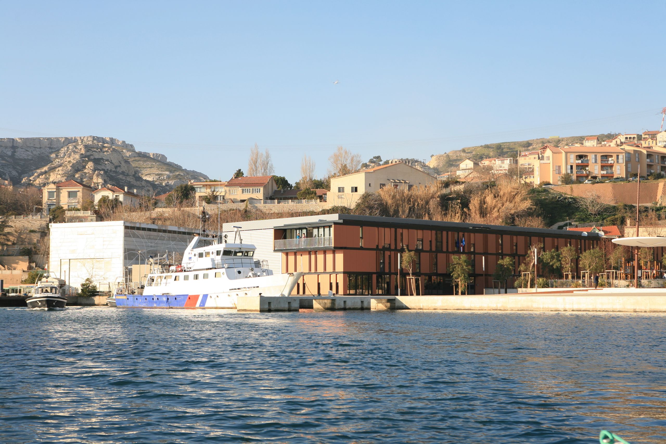 Vue d’ensemble du Département des Recherches Subaquatiques et Sous-Marines à Marseille, volume bas en bardage métallique brun sur quai, en front de mer avec navire accosté.