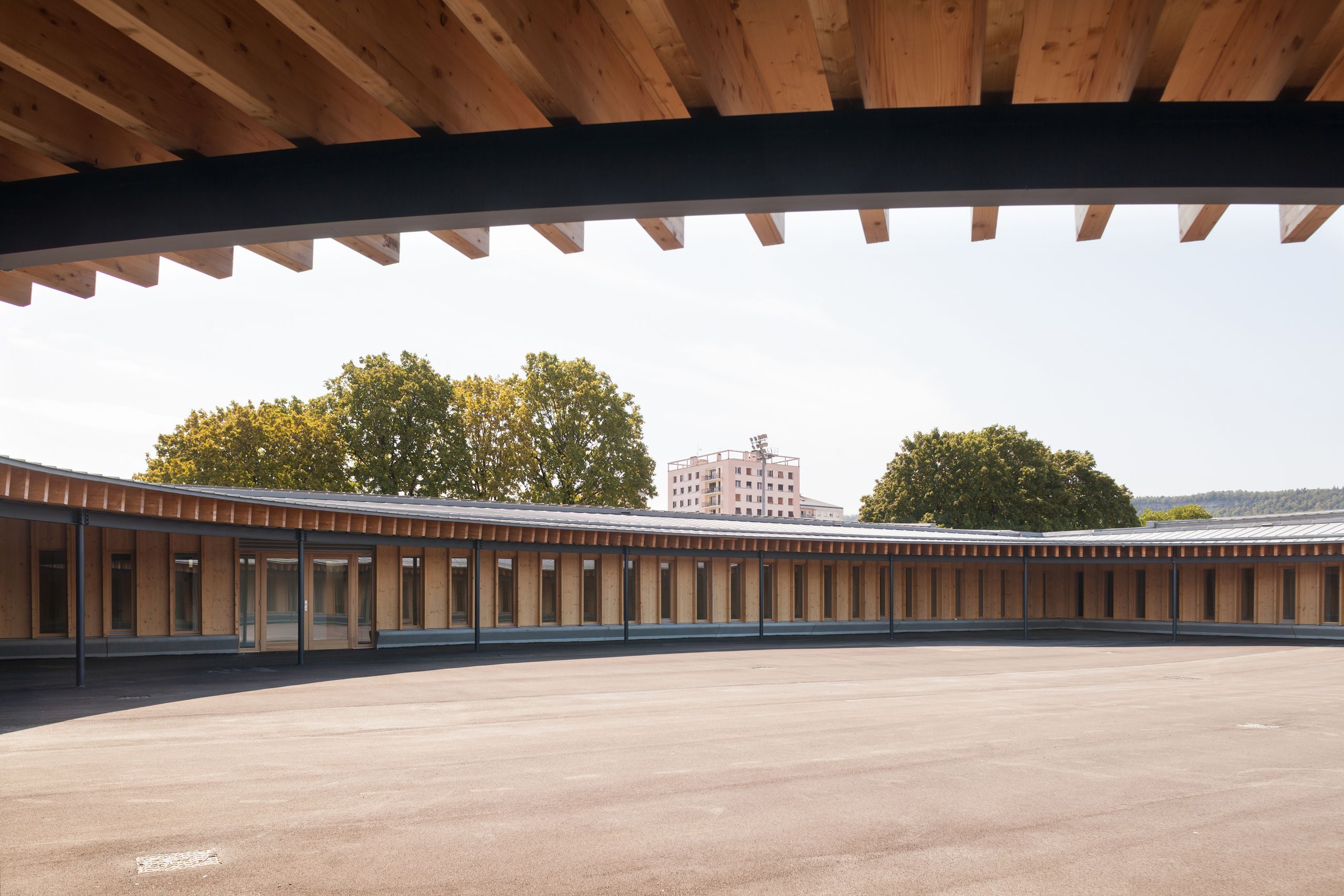 Cour elliptique de l’école élémentaire Hubert Reeves à Champagnole, entourée d’un volume bas en ossature bois et zinc, ouverte sur le paysage urbain en arrière-plan.