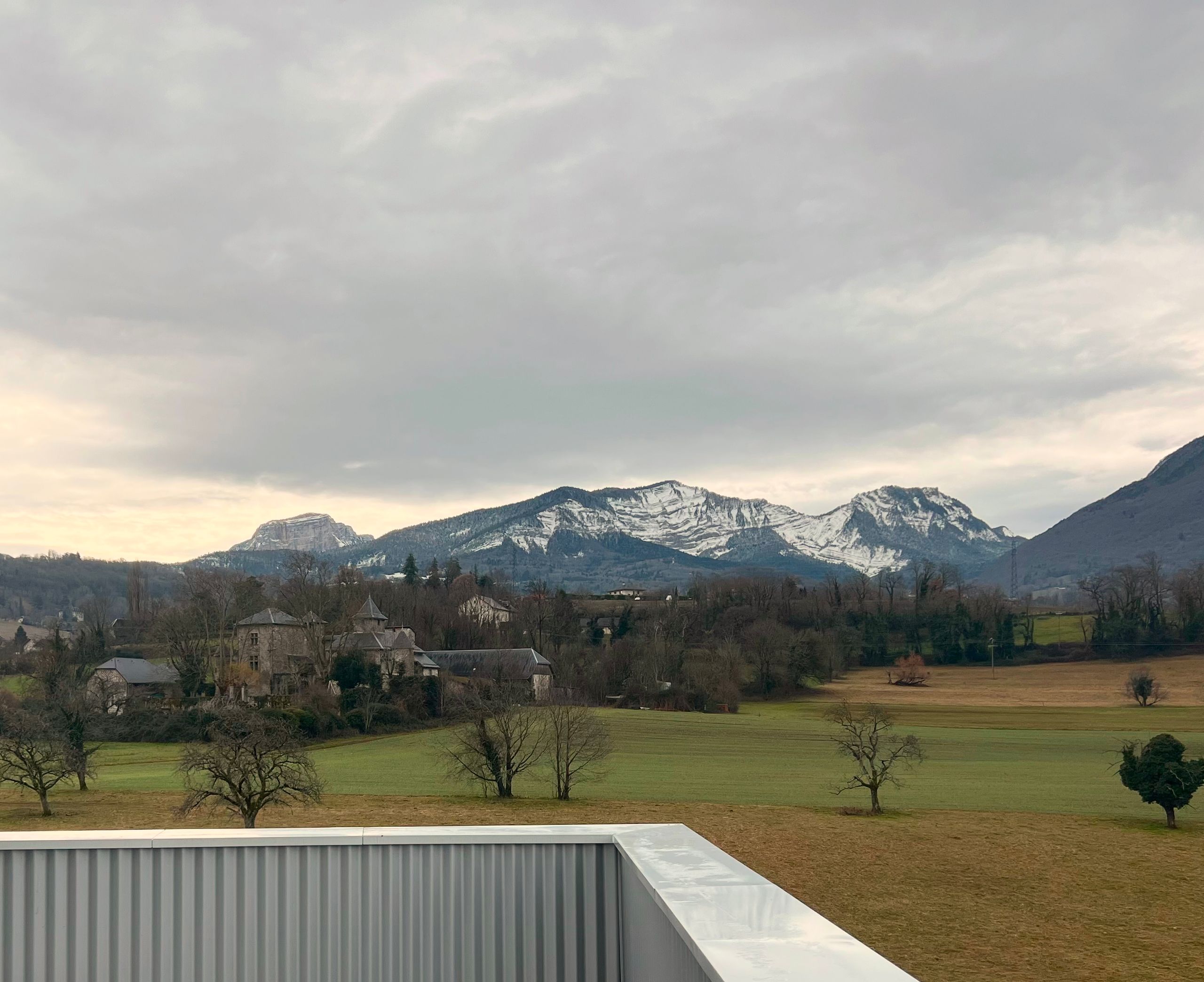 Vue depuis le projet de 142 logements à Cognin : acrotère métallique en premier plan, paysage rural et massif montagneux enneigé en arrière-plan.
