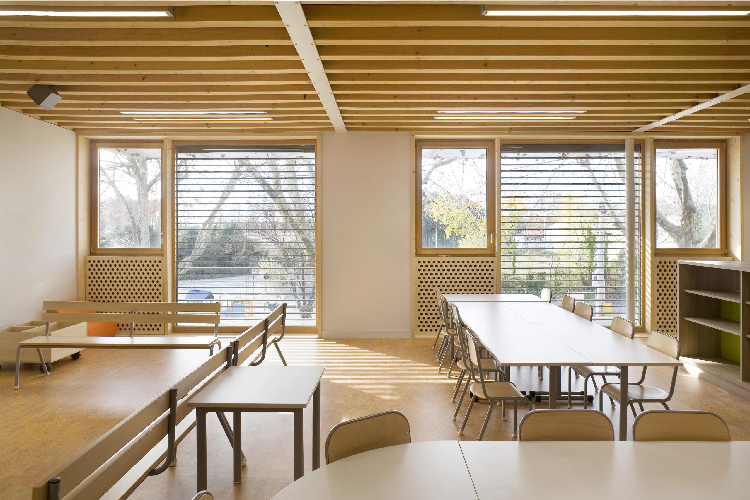 Intérieur de l’école Jean Carrière à Nîmes : salle de classe en ossature bois, plafond à poutres apparentes, grandes baies vitrées avec brise-soleil sur les platanes.