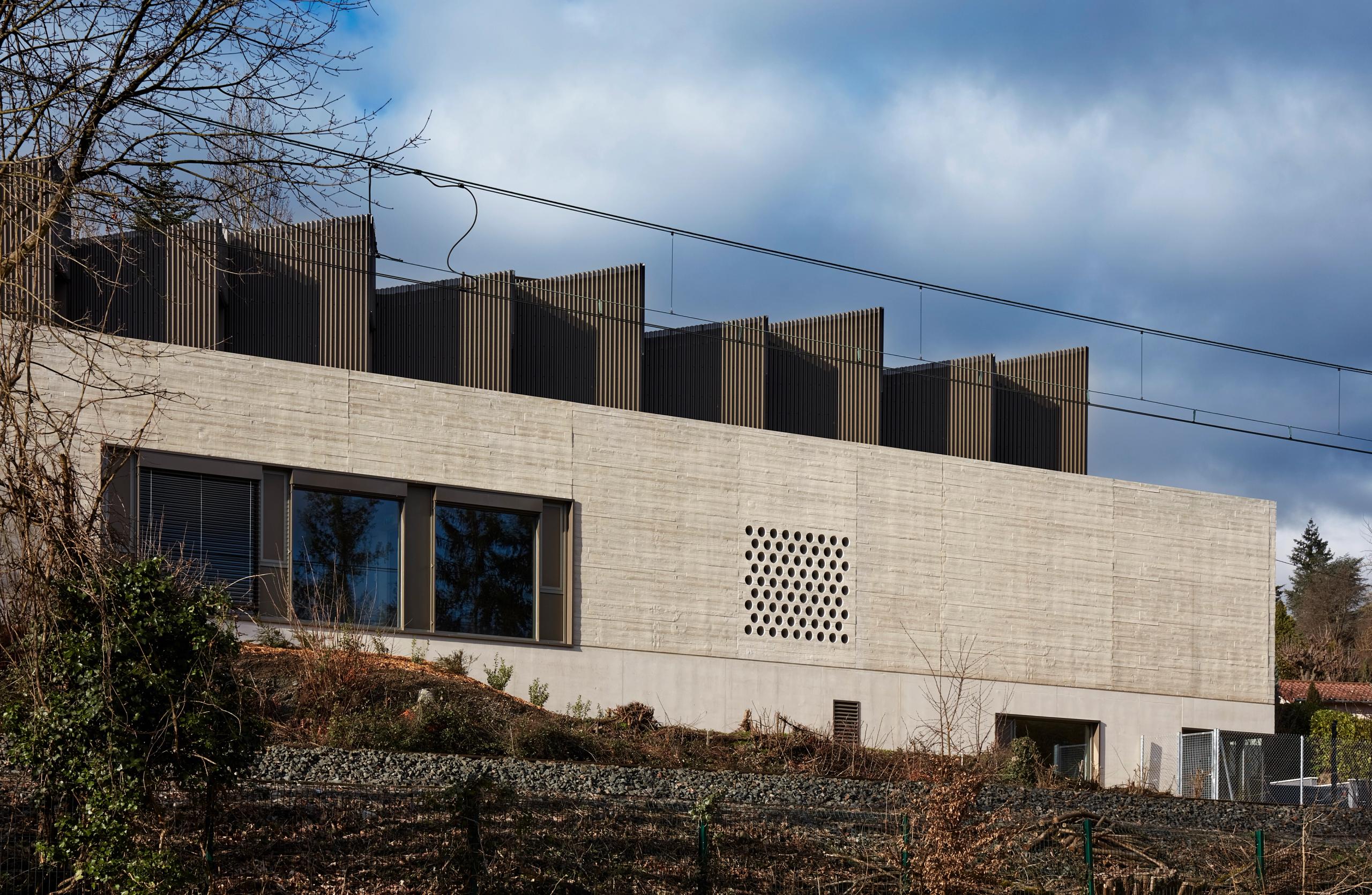 Façade de la Maison des Arts à Charbonnières-les-Bains, volume en béton strié sur socle enterré, couronnement bois-métal perforé le long de la voie ferrée.