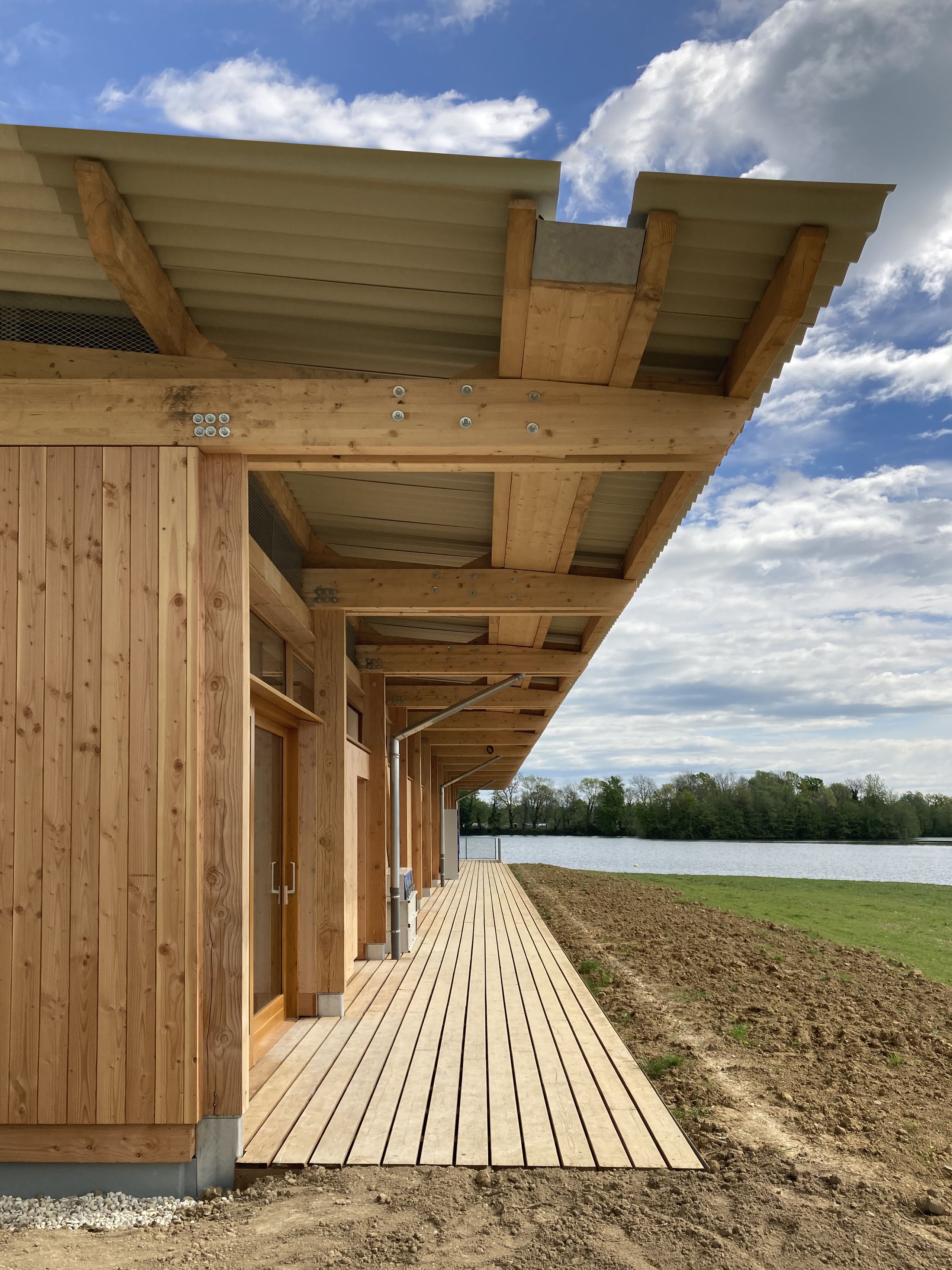 Maison des sports à Malafretaz : façade en ossature bois, auvent à toiture nervurée et coursive en lames de bois longeant le plan d’eau et le paysage ouvert.