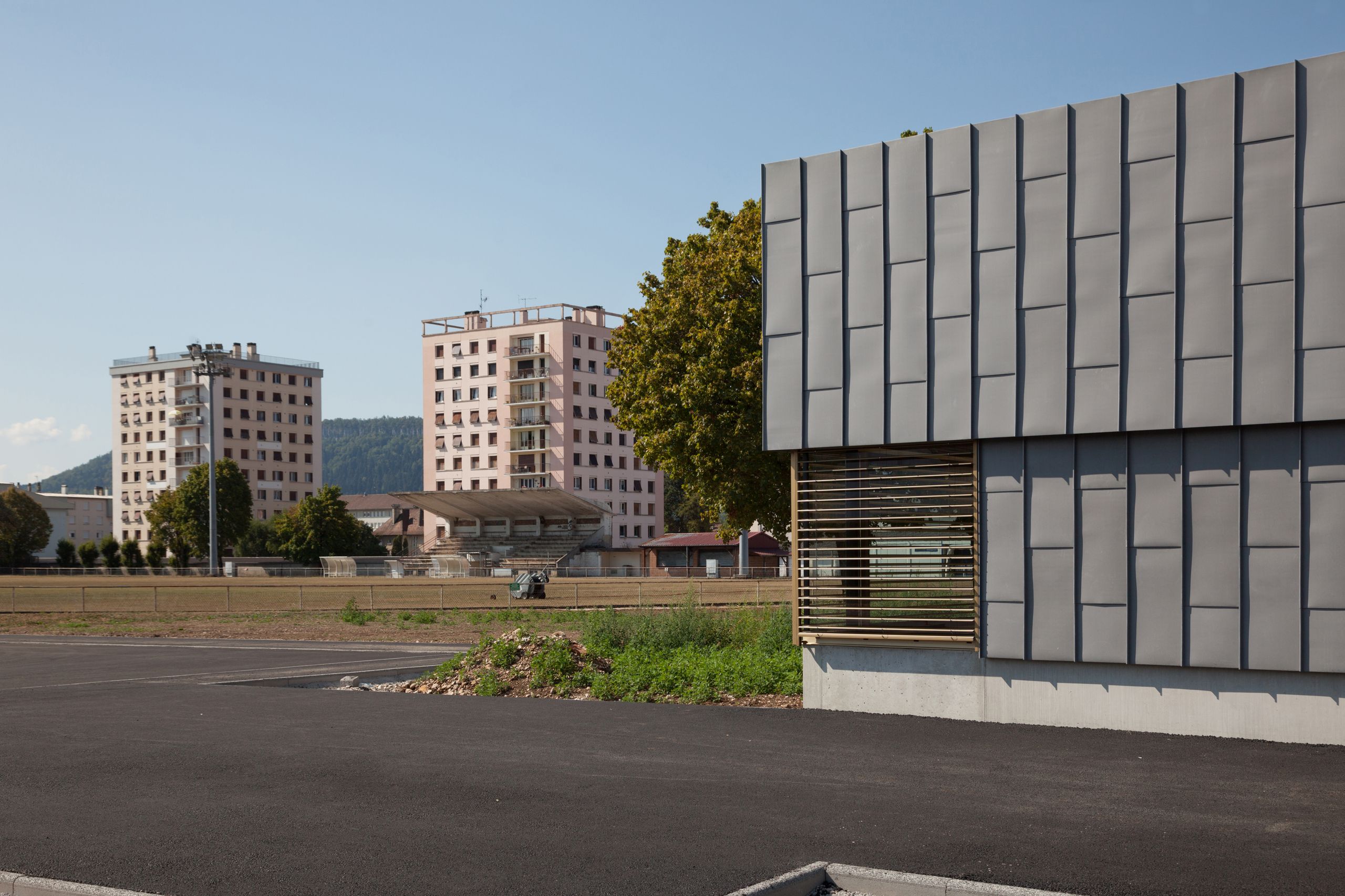 Façade en zinc de l’école élémentaire Hubert Reeves à Champagnole, percée d’une baie horizontale à lames bois, sur fond de tours d’habitation périurbaines.