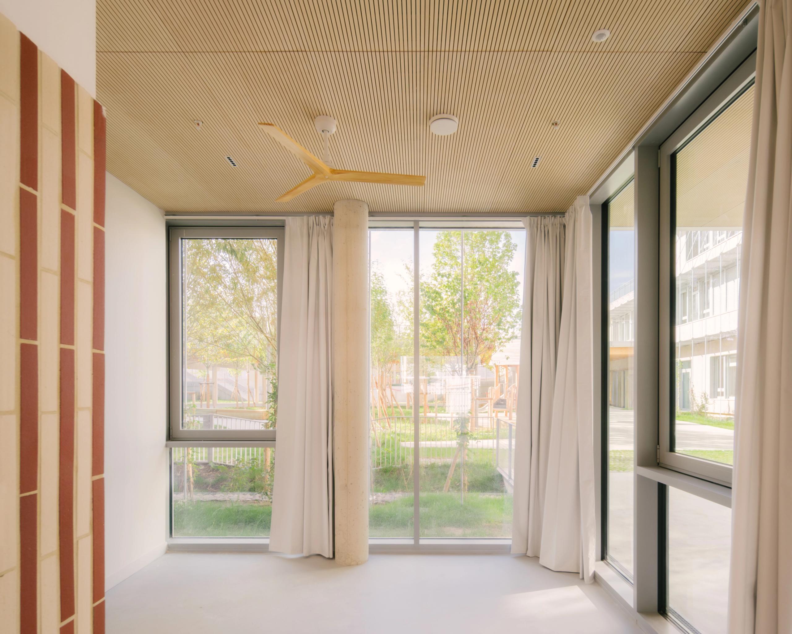 Intérieur du groupe scolaire Dominique Frelaut à Colombes : salle vitrée sur jardin, poteau béton, plafond bois rainuré et brise-soleil intégré.
