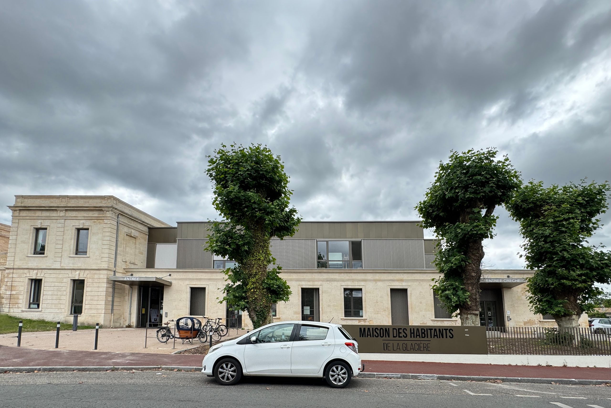 Vue de la maison des habitants restructurée à Mérignac, mêlant bâti en pierre existant et surélévation contemporaine en bardage métallique, sur rue arborée.
