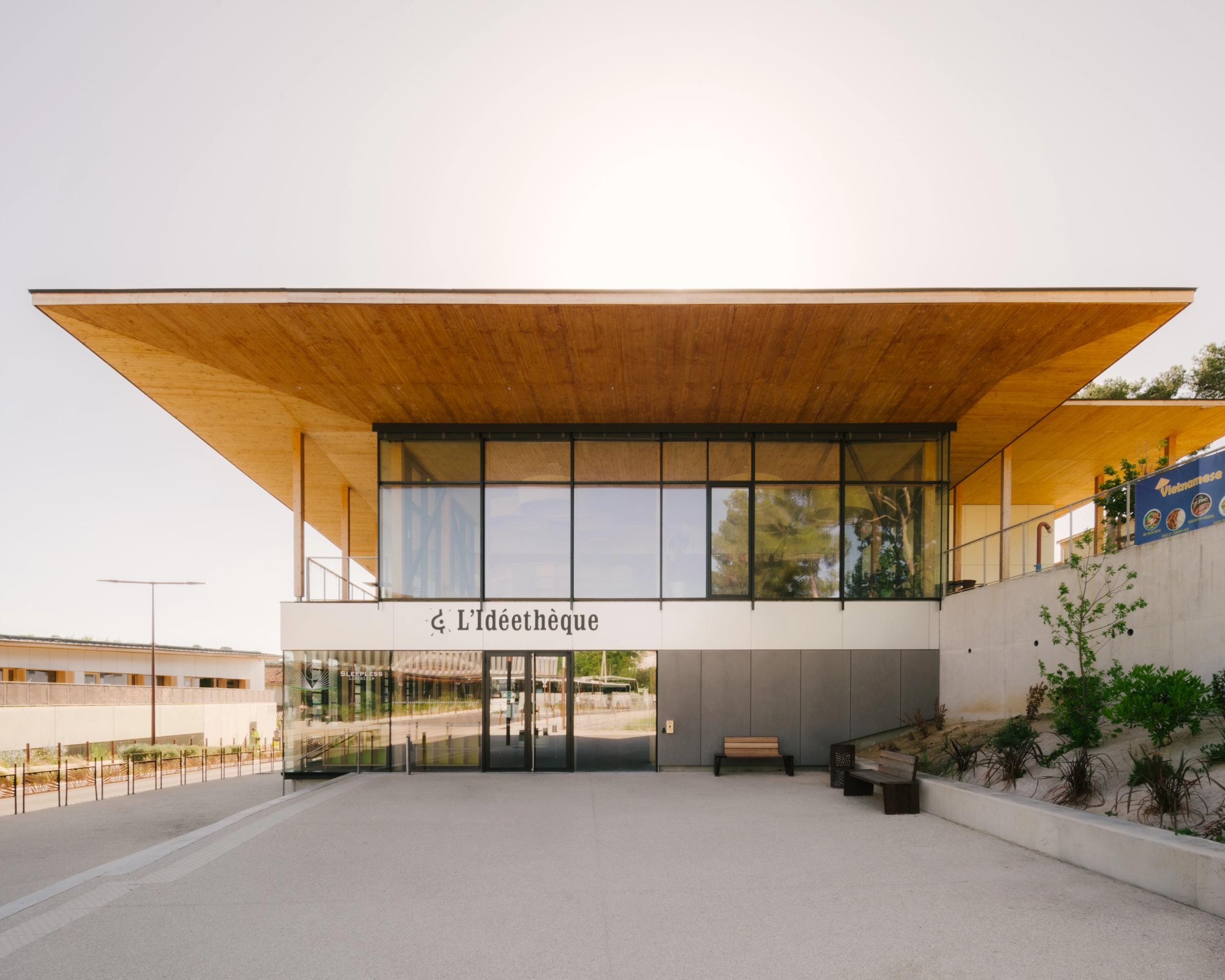 Façade de la médiathèque du groupe scolaire Claudie Haigneré aux Pennes-Mirabeau, volume vitré sous large toiture bois débordante, parvis minéral en pente.