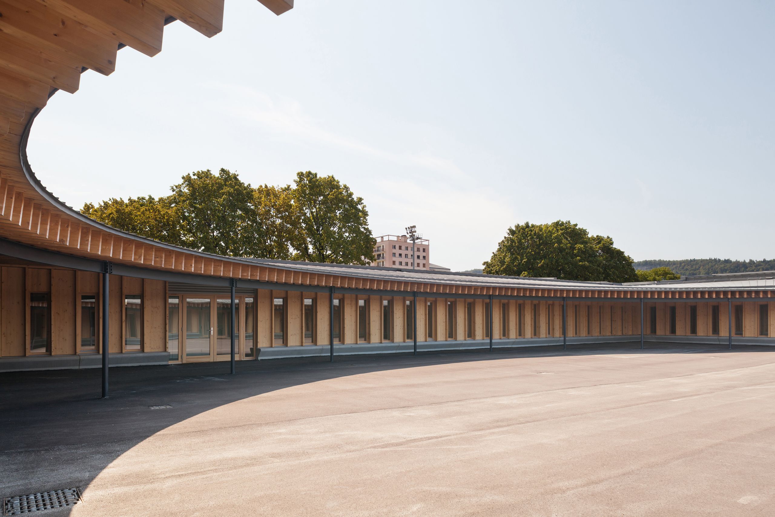 Cour elliptique de l’école élémentaire Hubert Reeves à Champagnole, entourée d’une galerie en ossature bois apparente sous toiture courbe en zinc.