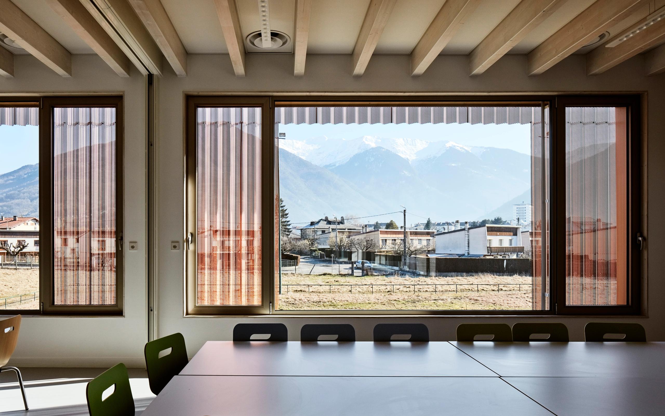 Interior of Simone Veil Early Childhood Center in Albertville, with large framed windows, perforated metal screens and Alpine mountains visible beyond.