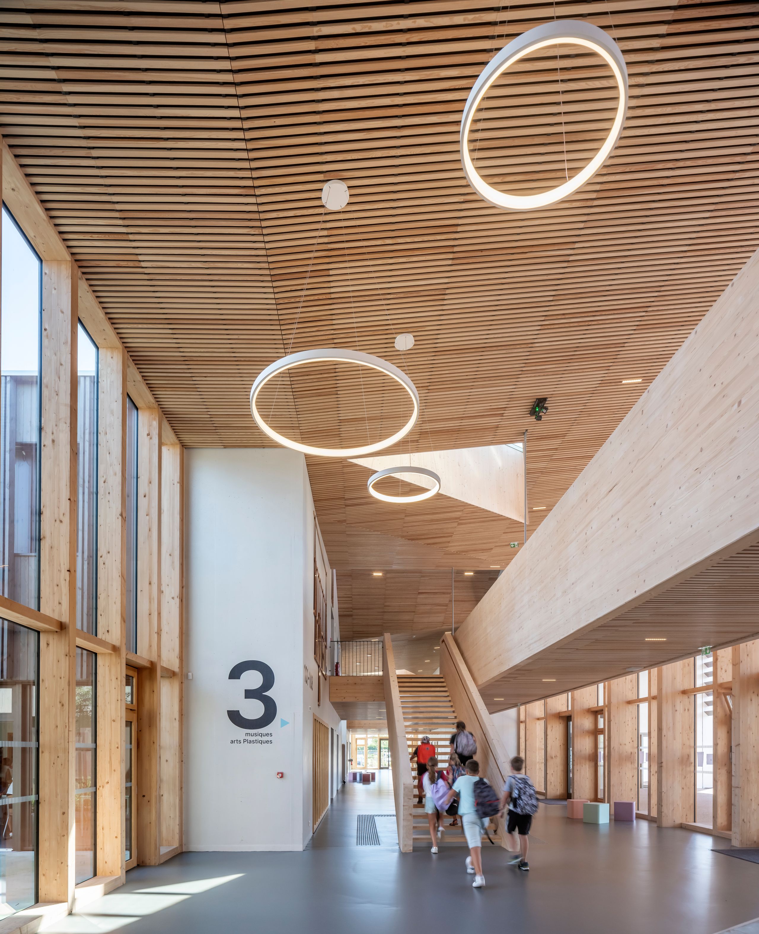 Rue intérieure du Collège du Ban à Vagney, structure bois apparent, double hauteur vitrée, escalier central et luminaires circulaires suspendus.