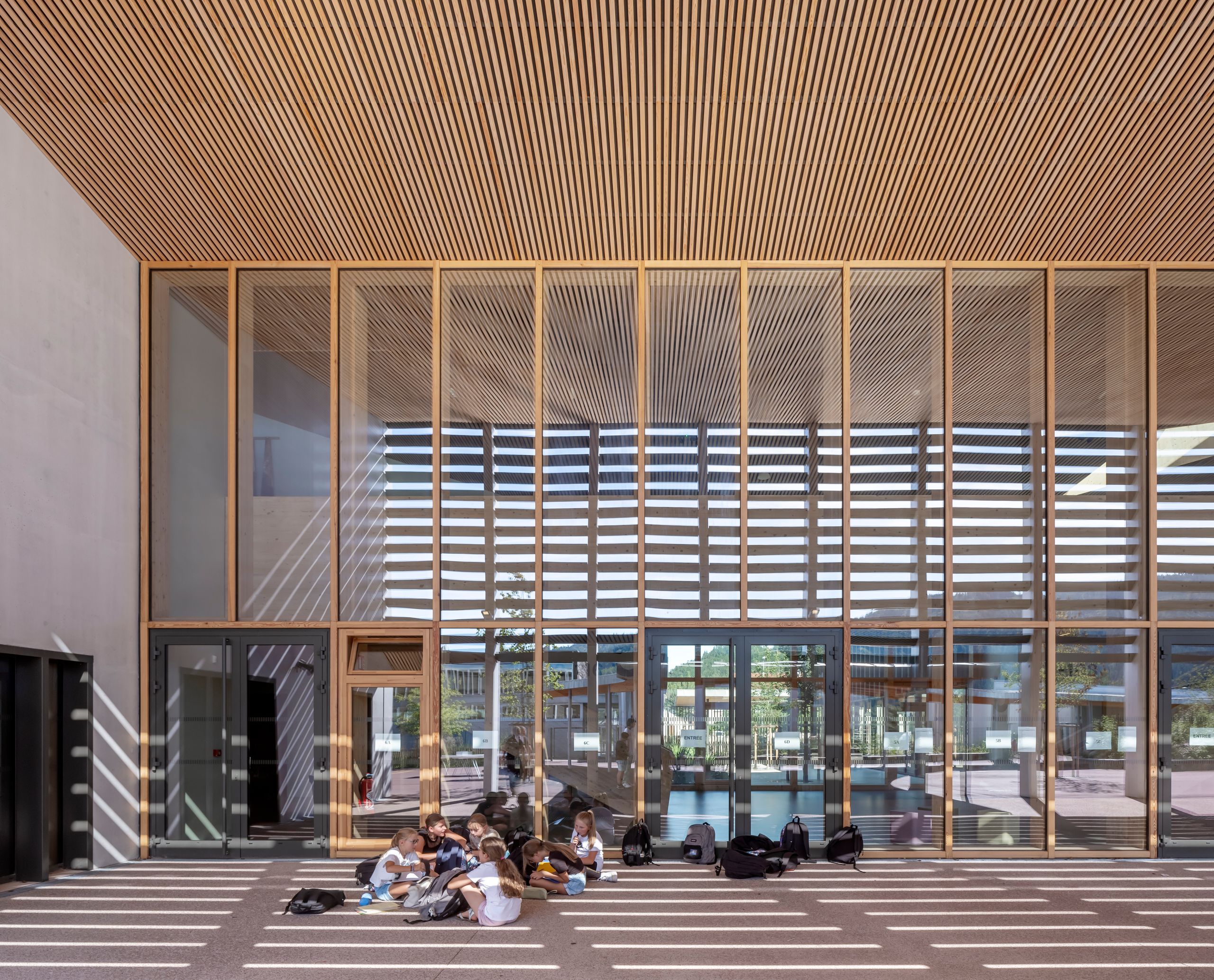 Vue du portique d’entrée du Collège du Ban à Vagney : une façade largement vitrée en double hauteur, rythmée par une ossature bois verticale et un plafond à lames de bois continues, crée un préau couvert où la lumière forme des bandes horizontales au sol ; des élèves assis sous l’auvent illustrent l’usage de cette rue intérieure comme espace de transition entre cour et bâtiment.