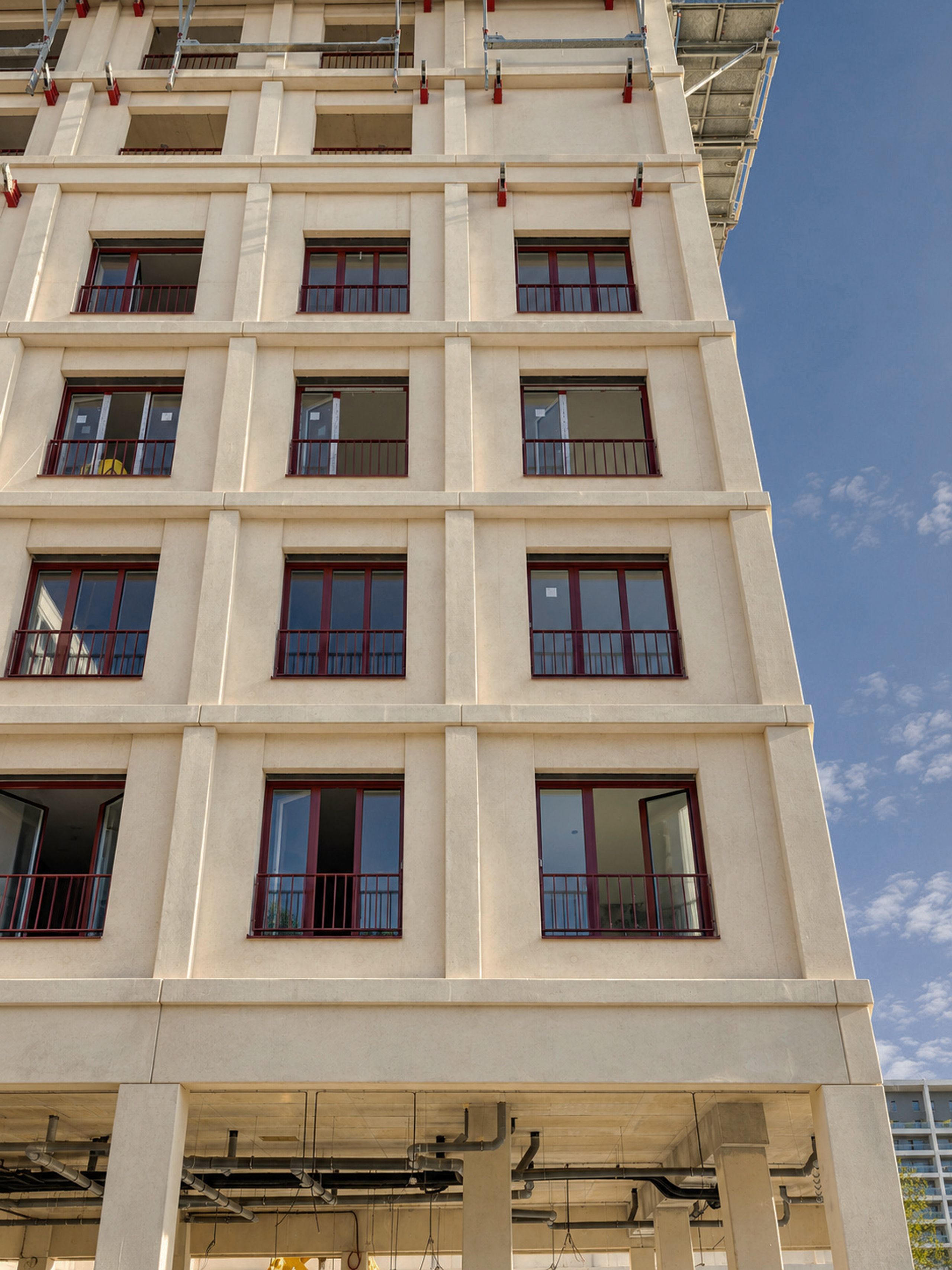 À Bordeaux, façade préfabriquée tramée de l’îlot 8.10, avec baies et garde-corps rouges ; rez-de-chaussée sur poteaux et échafaudages en toiture.