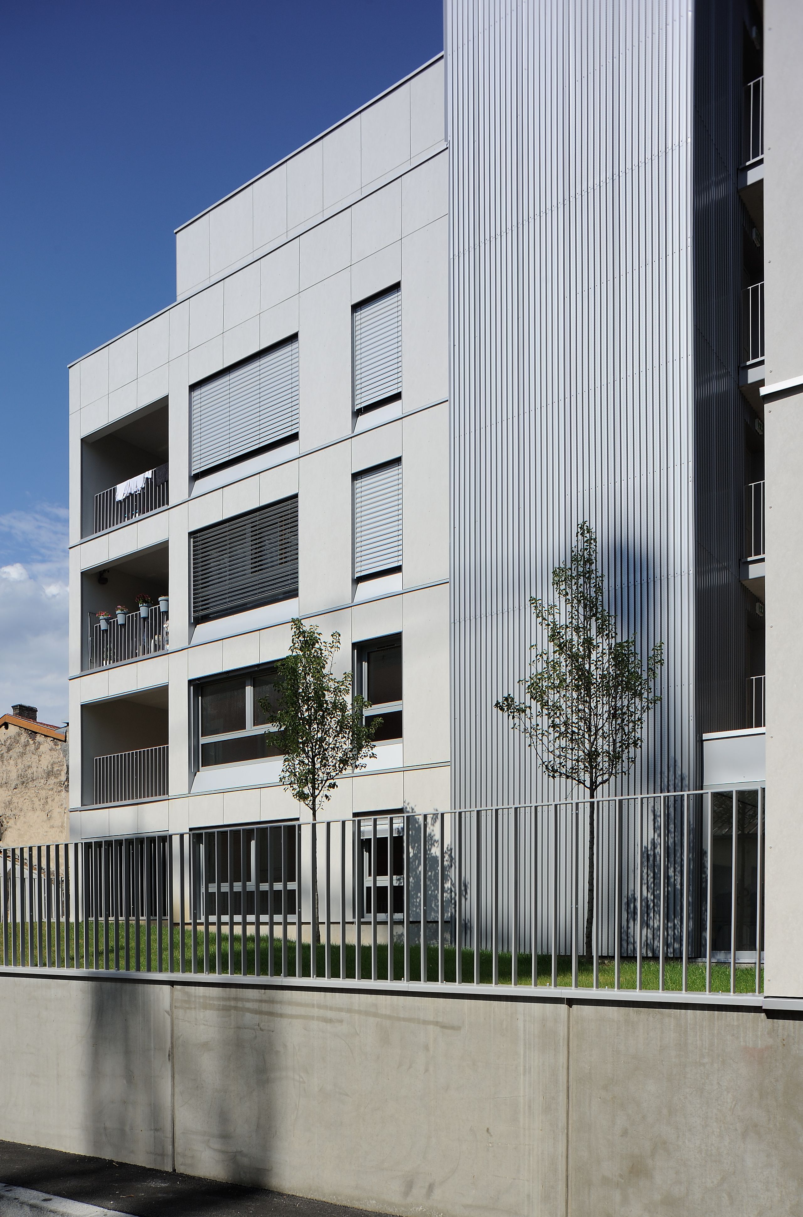 Façade des 28 logements Tectoniques à Villeurbanne, volumes fractionnés, loggias en retrait, bardage métallique vertical et clôture sur jardin surélevé.