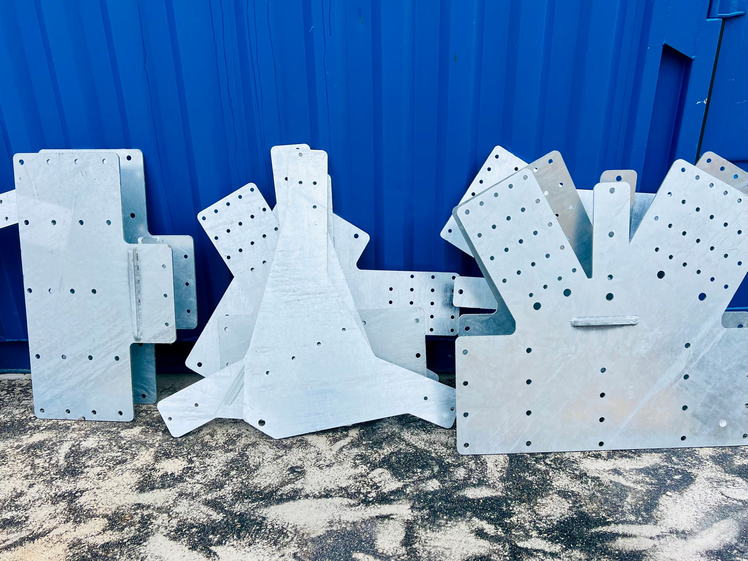 Platines métalliques de contreventement pour la structure bois du Groupe scolaire Louis Pergaud à Levier, posées au sol devant une paroi bleue.