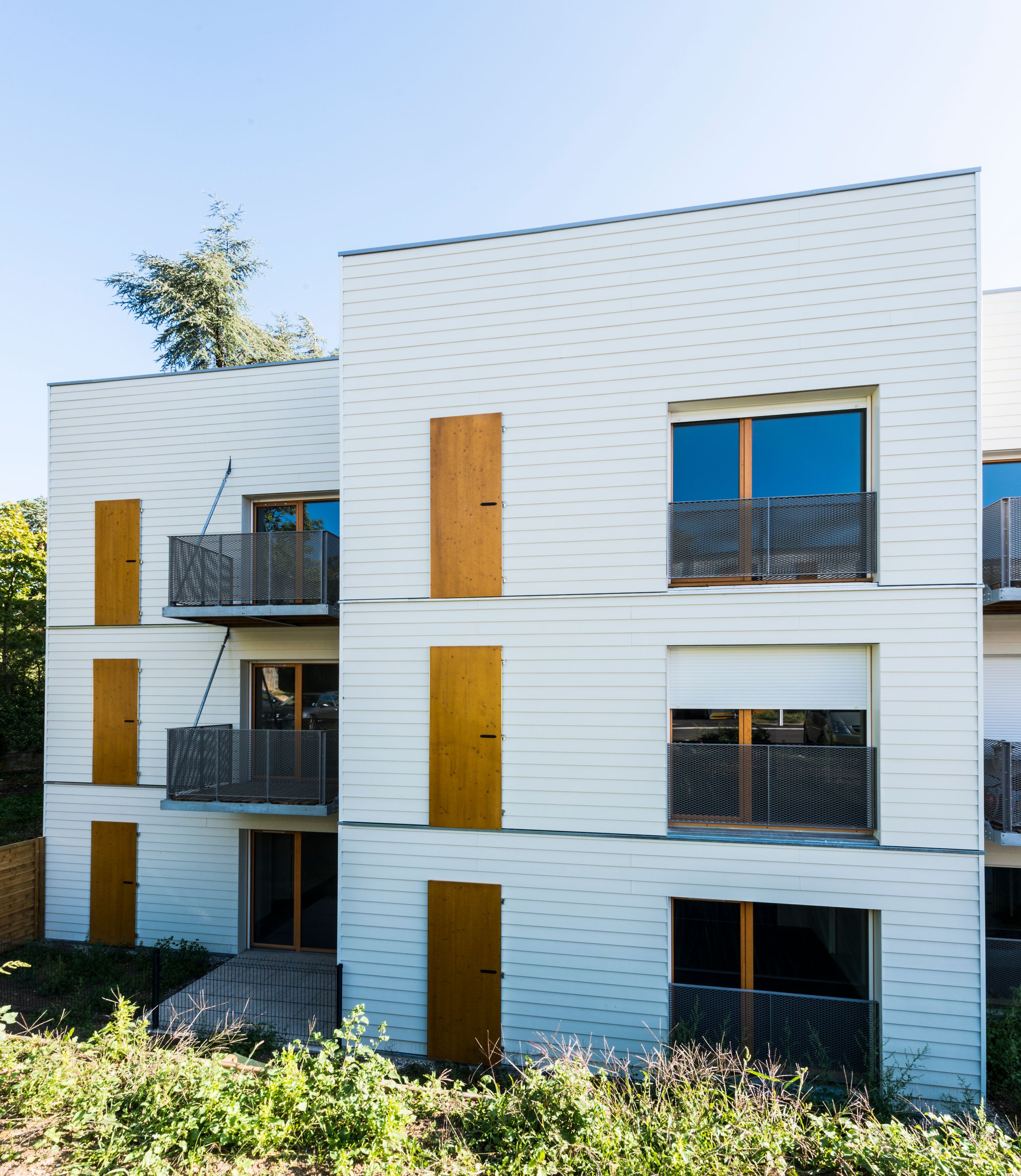 Façade de logements Tectoniques à Rive-de-Gier : volumes fragmentés en bardage clair, menuiseries bois, balcons métalliques et implantation sur terrain en pente.
