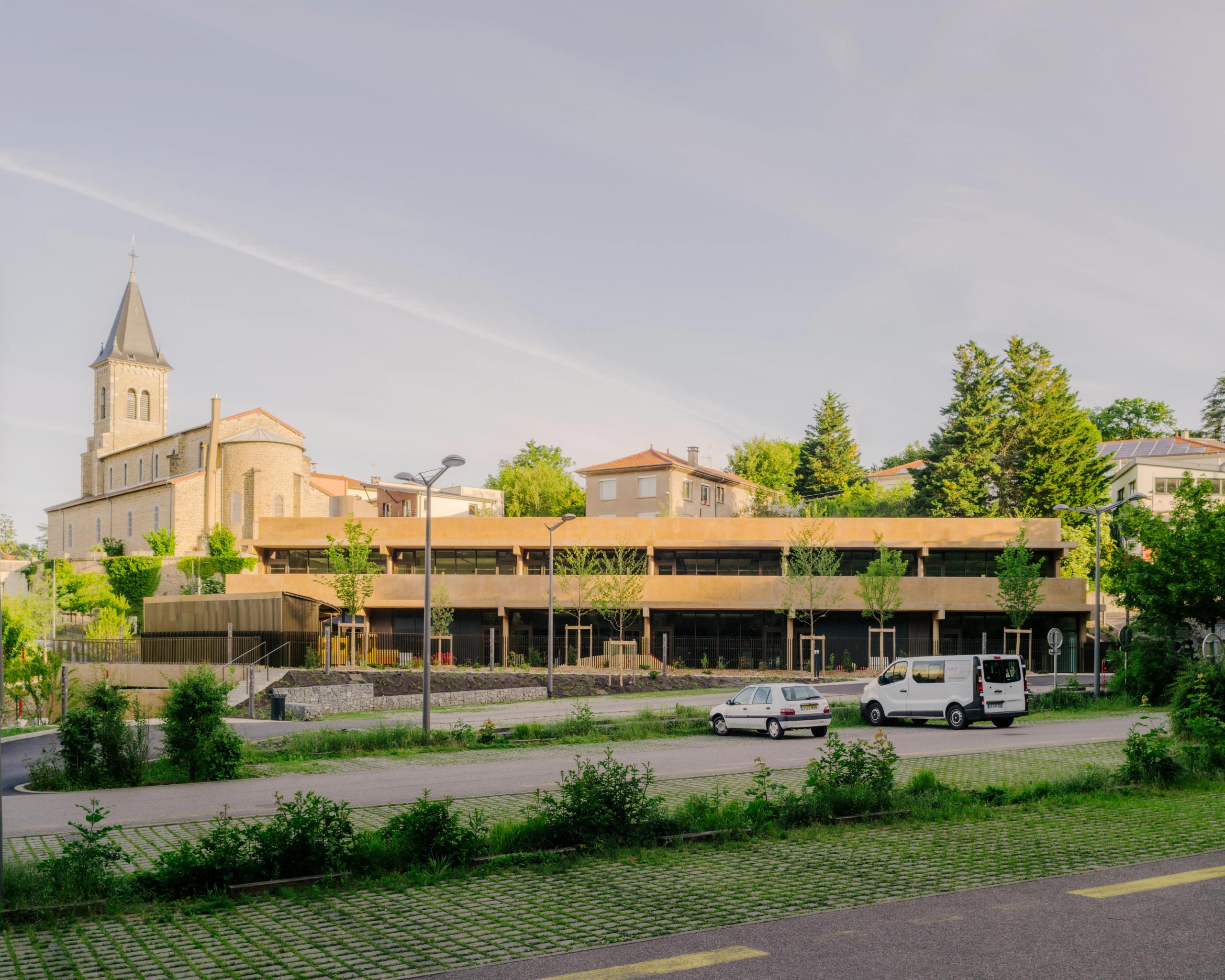 Vue de la Maison des familles à Limonest : volume tellurique en terrasses de béton teinté, façades horizontales vitrées, inséré sous le parvis de l’église.