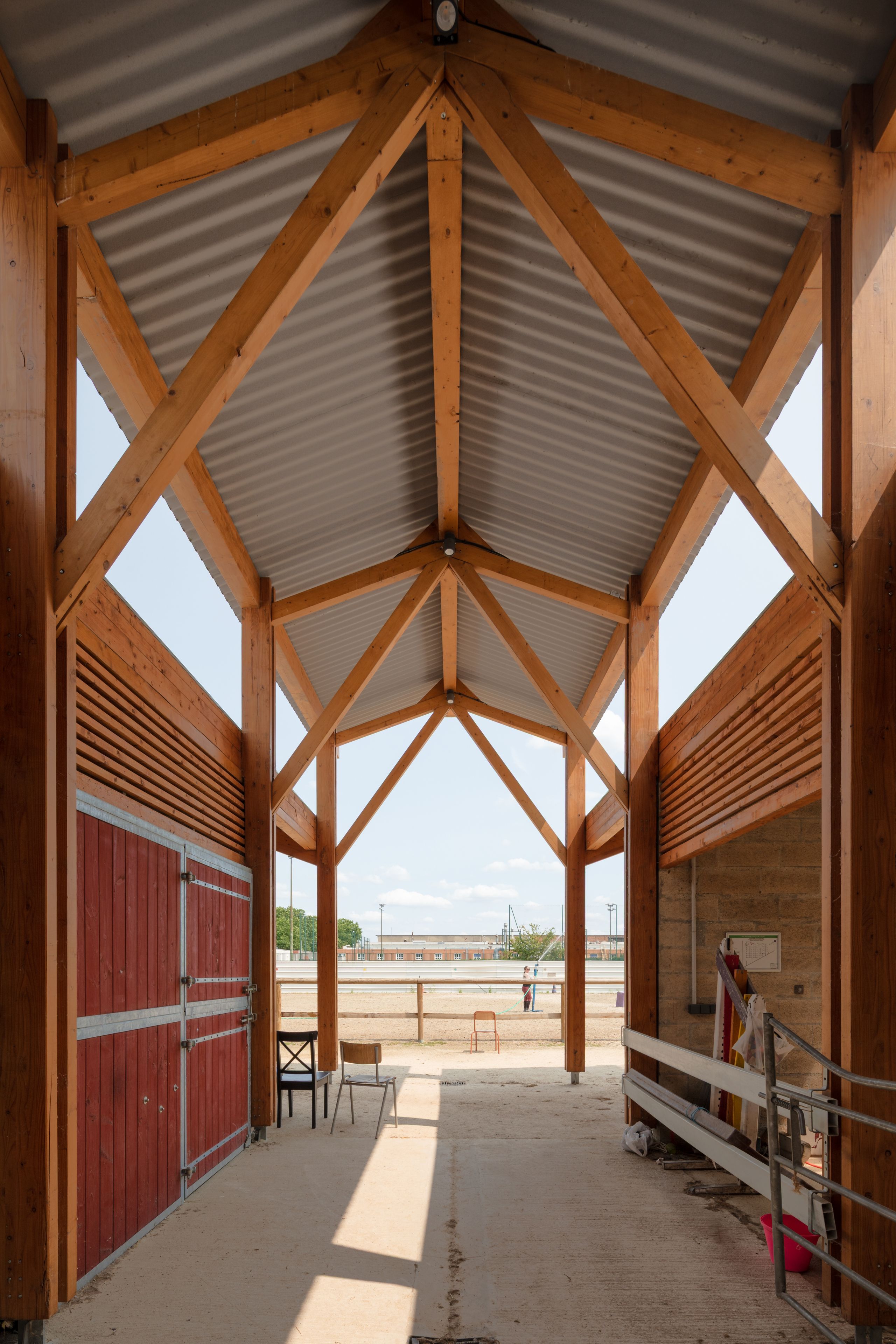 Gennevilliers, centre équestre Tectoniques : passage couvert en structure bois apparente, toiture ondulée, bardage rouge et vues cadrées sur la carrière.