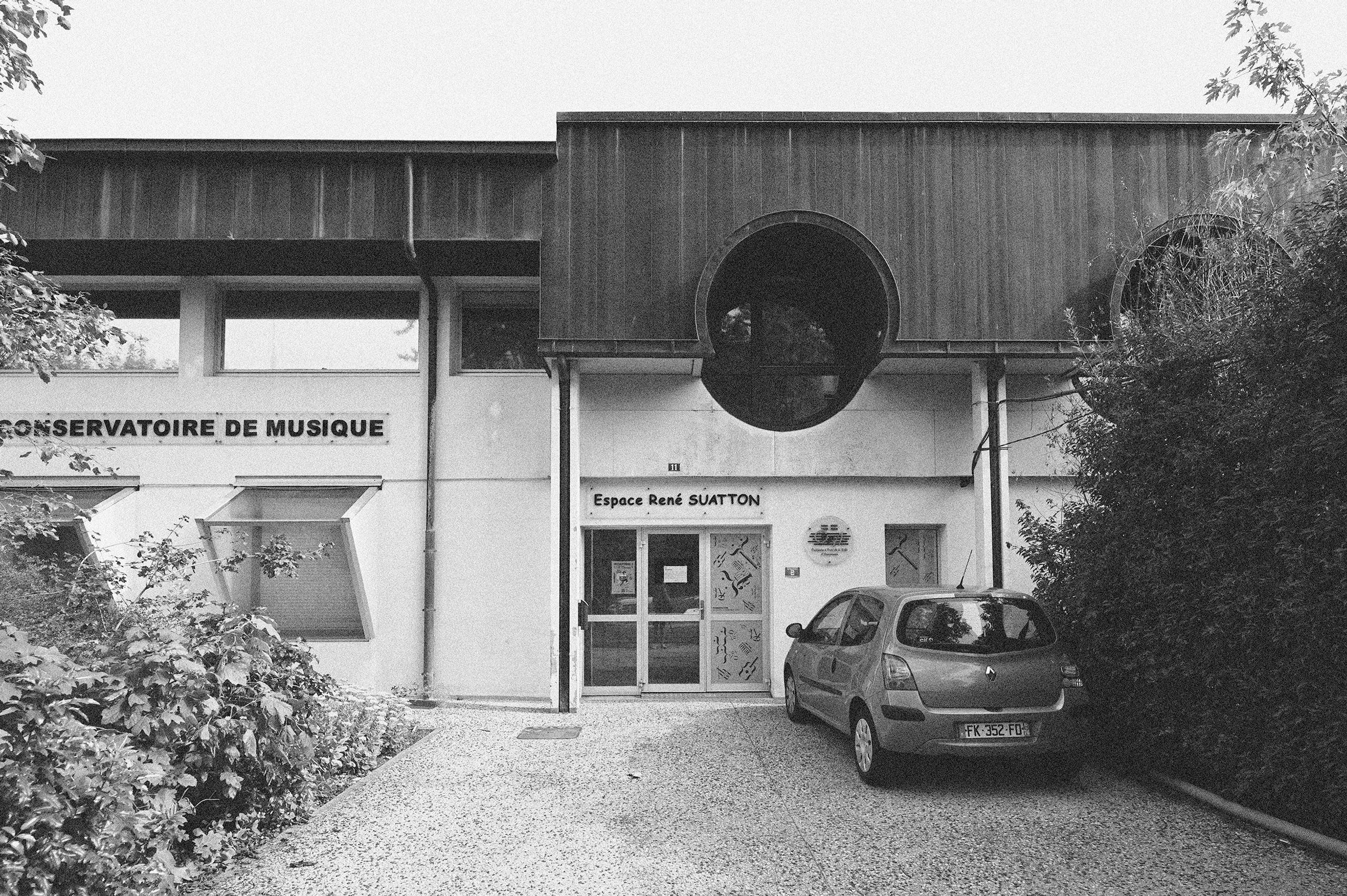 État existant du conservatoire d’Annemasse : façade en béton, bardage sombre et baie ronde signalant l’entrée, bordée par la végétation.