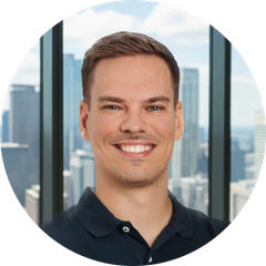 Lächelnder Mann mit kurzen braunen Haaren in dunklem Polo, vor großen Fenstern mit Blick auf die Stadt.