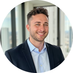 Lächelnder Mann mit lockigem Haar und Bart in dunklem Sakko und hellblauem Hemd vor großen Fenstern.