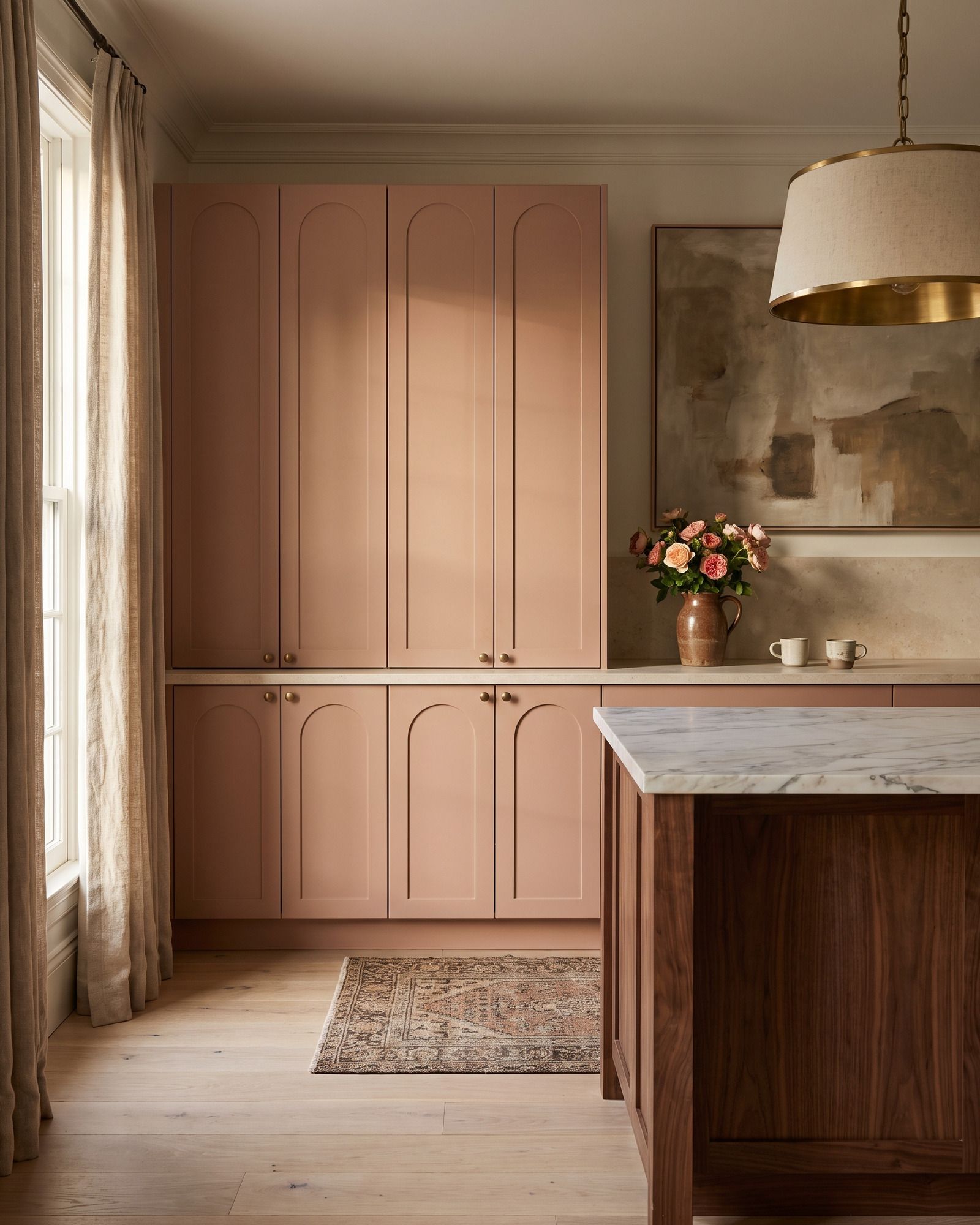 A kitchen with a tall run of frameless doors painted clay pink, each with a recessed panel arched at the top, small brass knobs, a limestone counter and a walnut island