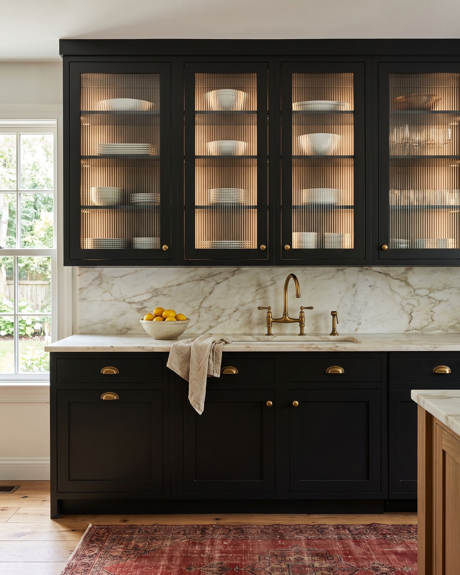 A kitchen with soft-black upper cabinets whose doors hold bold ribbed glass, the plates behind streaked into vertical lines, lit inside, over a Calacatta backsplash and a brass bridge faucet