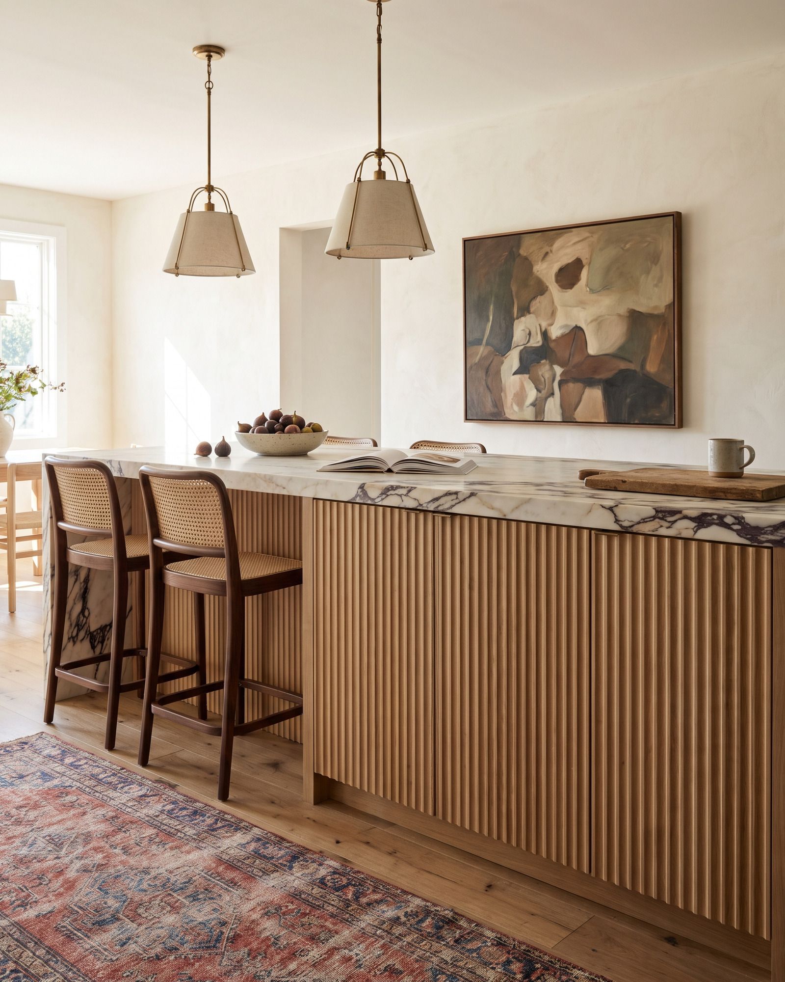 A kitchen island clad in fluted white oak doors with vertical concave grooves, under a thick honed Calacatta Viola top, cane counter stools and linen pendants