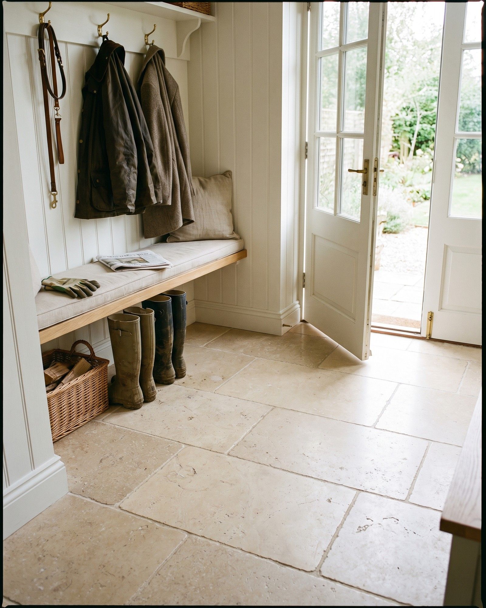 A mudroom with a floor of large honed beige limestone slabs, an oak bench, coats on hooks and boots by an open garden door