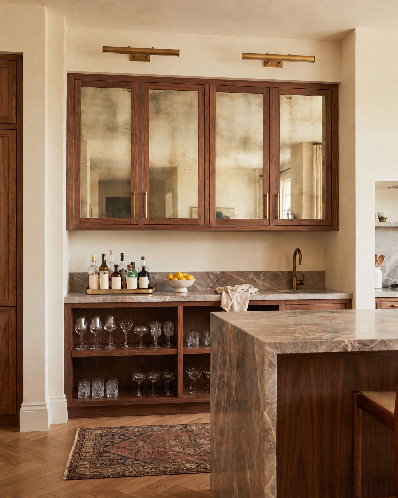 A bar with walnut upper cabinets whose doors hold foxed antique mirror, brass picture lights above, a brown marble counter with bottles and glasses