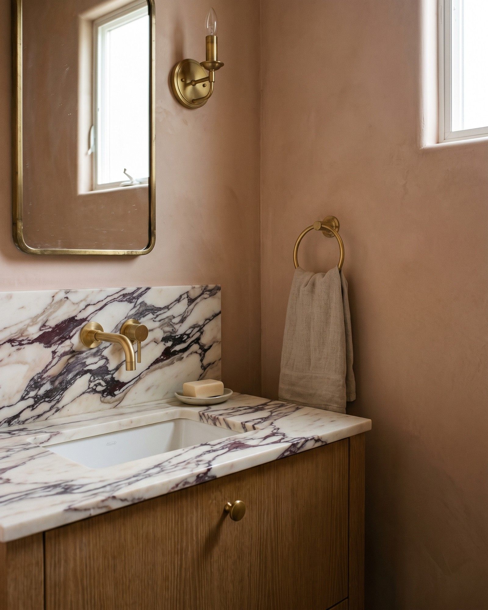A powder room vanity in honed Calacatta Viola marble, burgundy and violet veins on cream, on a rift oak base against clay-pink plaster