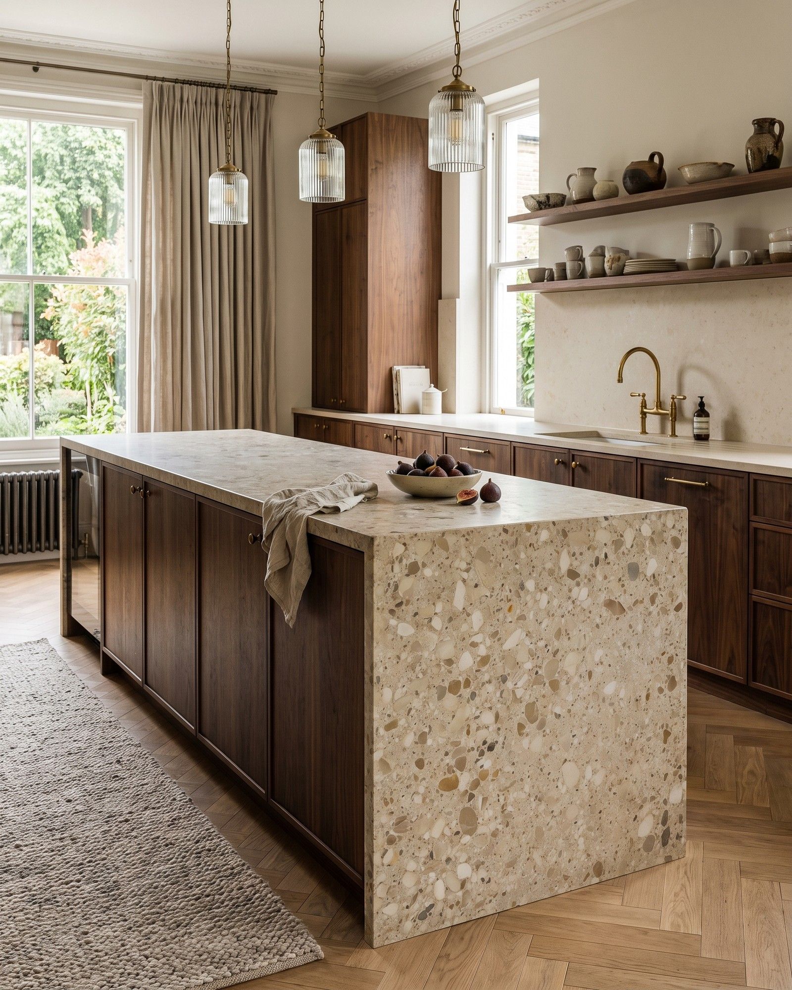 A kitchen island with a thick top and waterfall end in honed Ceppo di Gré, a pebbled beige conglomerate, on a dark walnut base over a herringbone oak floor