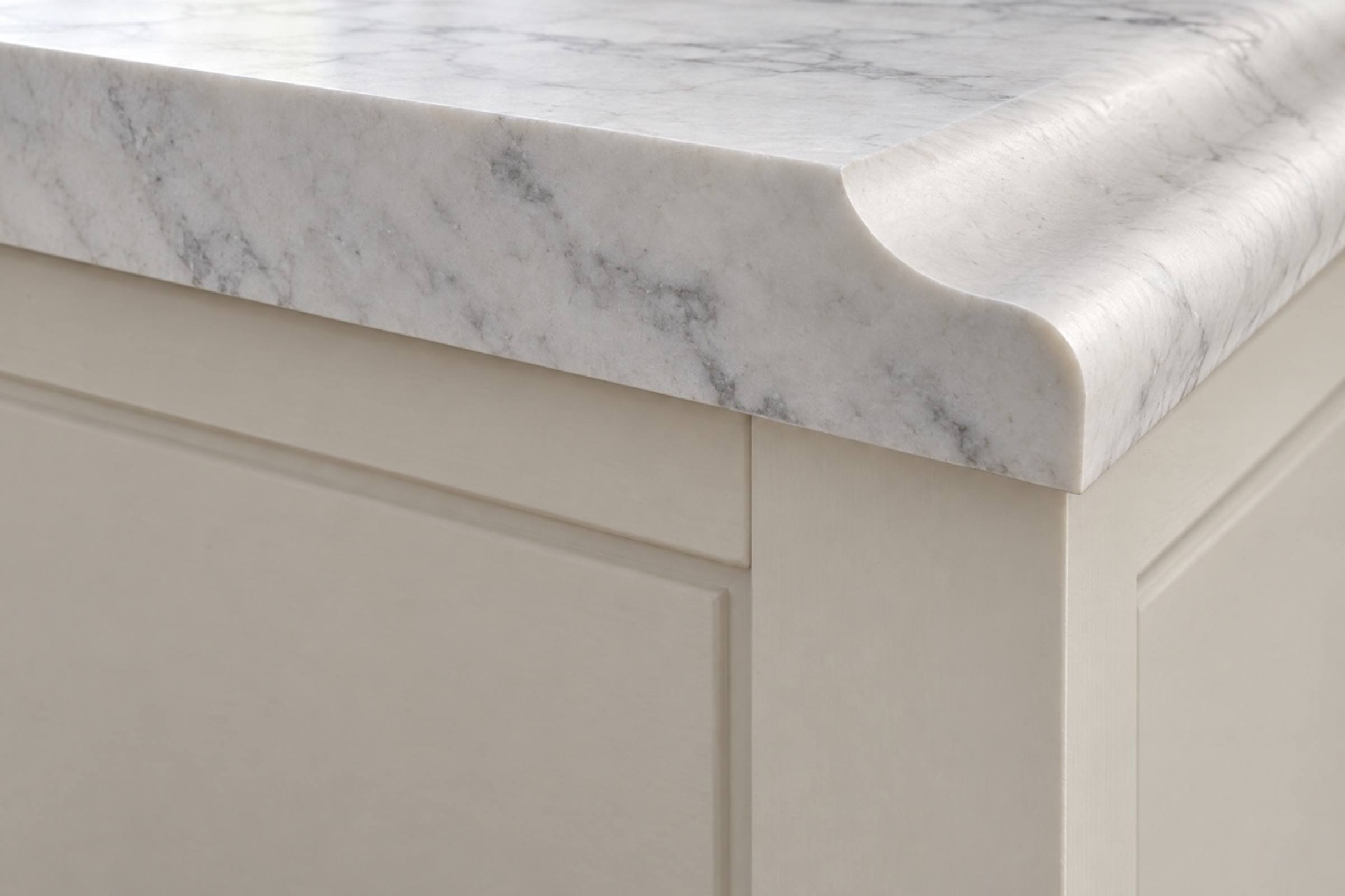 The cut end of a white marble countertop showing an ogee edge in section, over cream cabinets