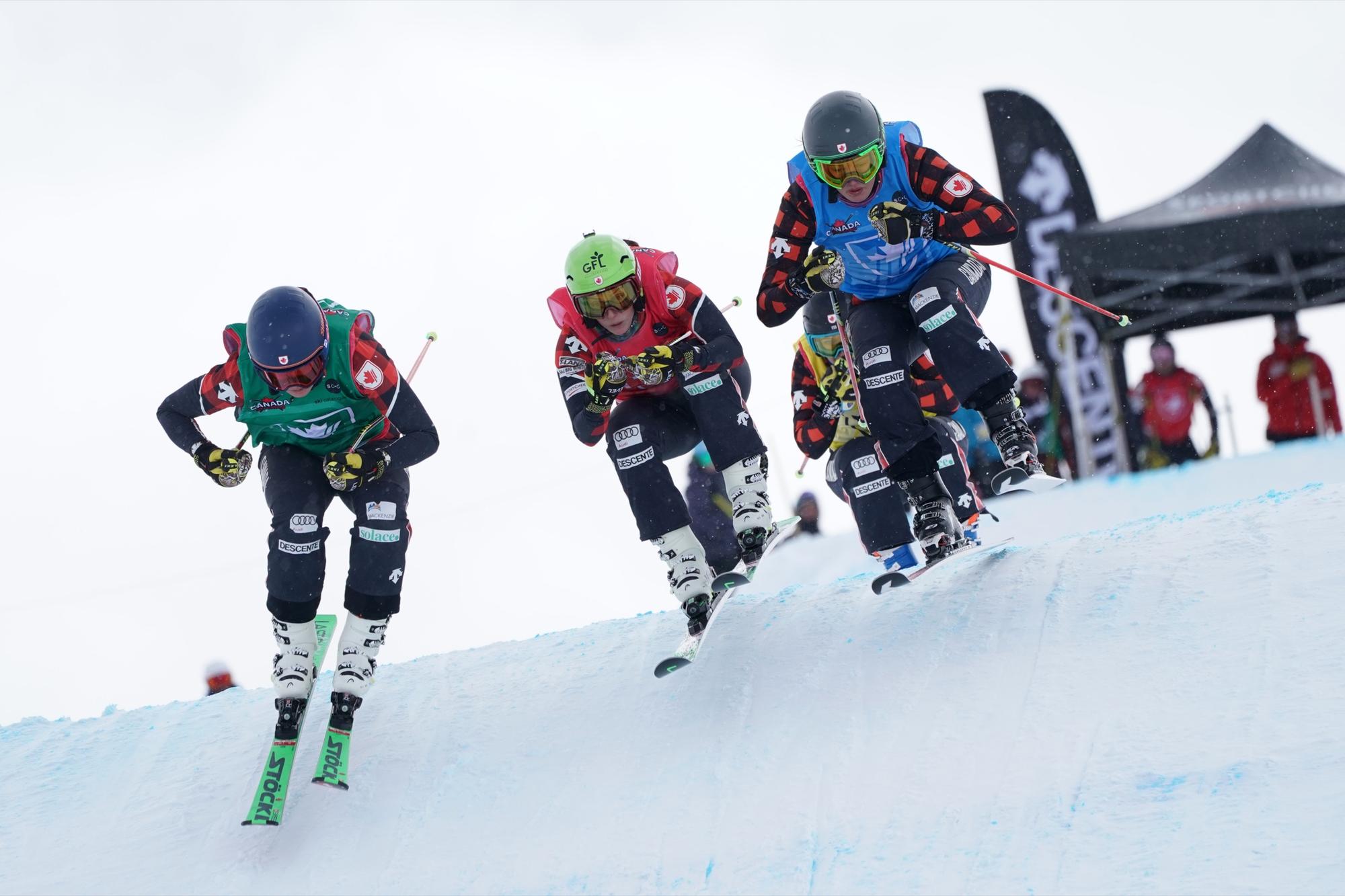 Three ski cross racers in Canada bibs airborne over a roller on a race course