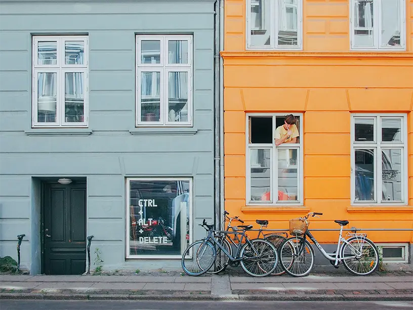Farverige byejendomme med cykler parkeret langs facaden og kvinde i vindue
