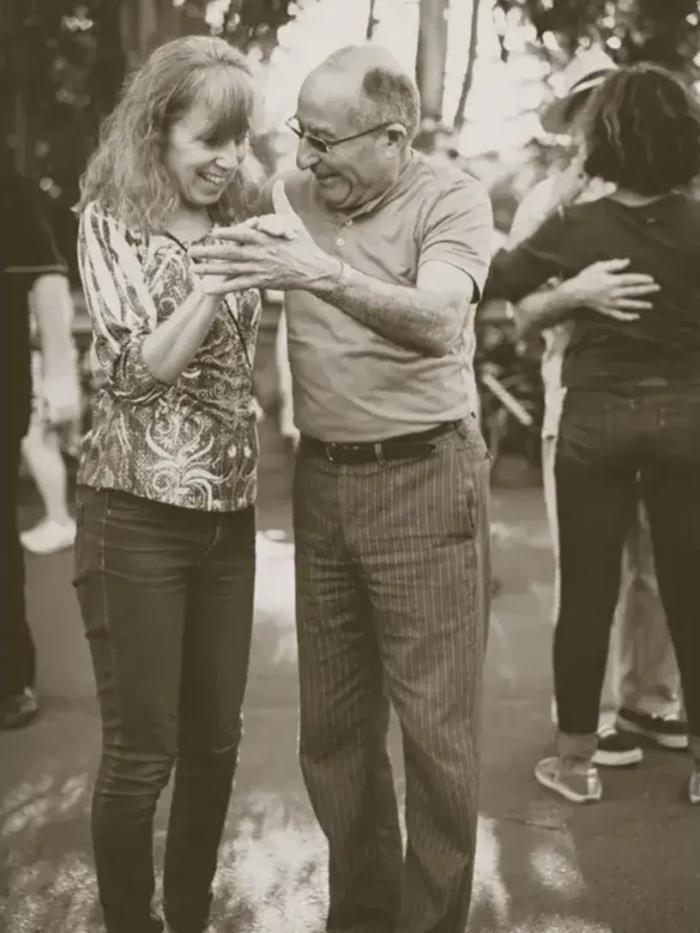 Ældre mand og yngre kvinde danser smilende sammen udendørs.