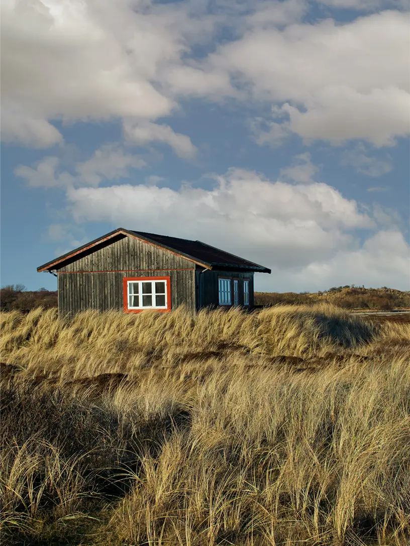Lille træhus i klitterne omgivet af marehalm under en skyet himmel.