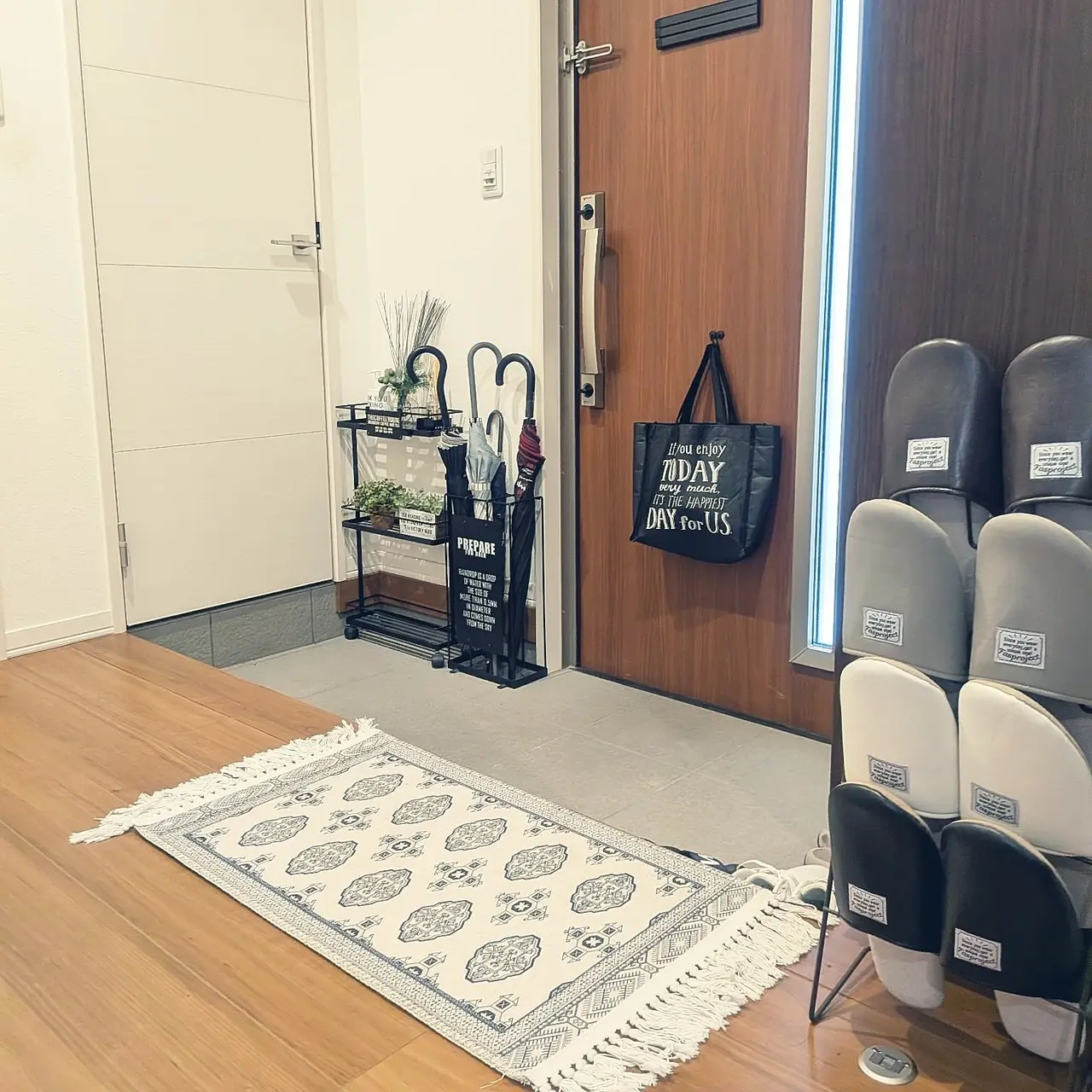 A tidy small Japanese entryway with a slim umbrella rack by the front door, a narrow rug, and slippers stored neatly to the side.