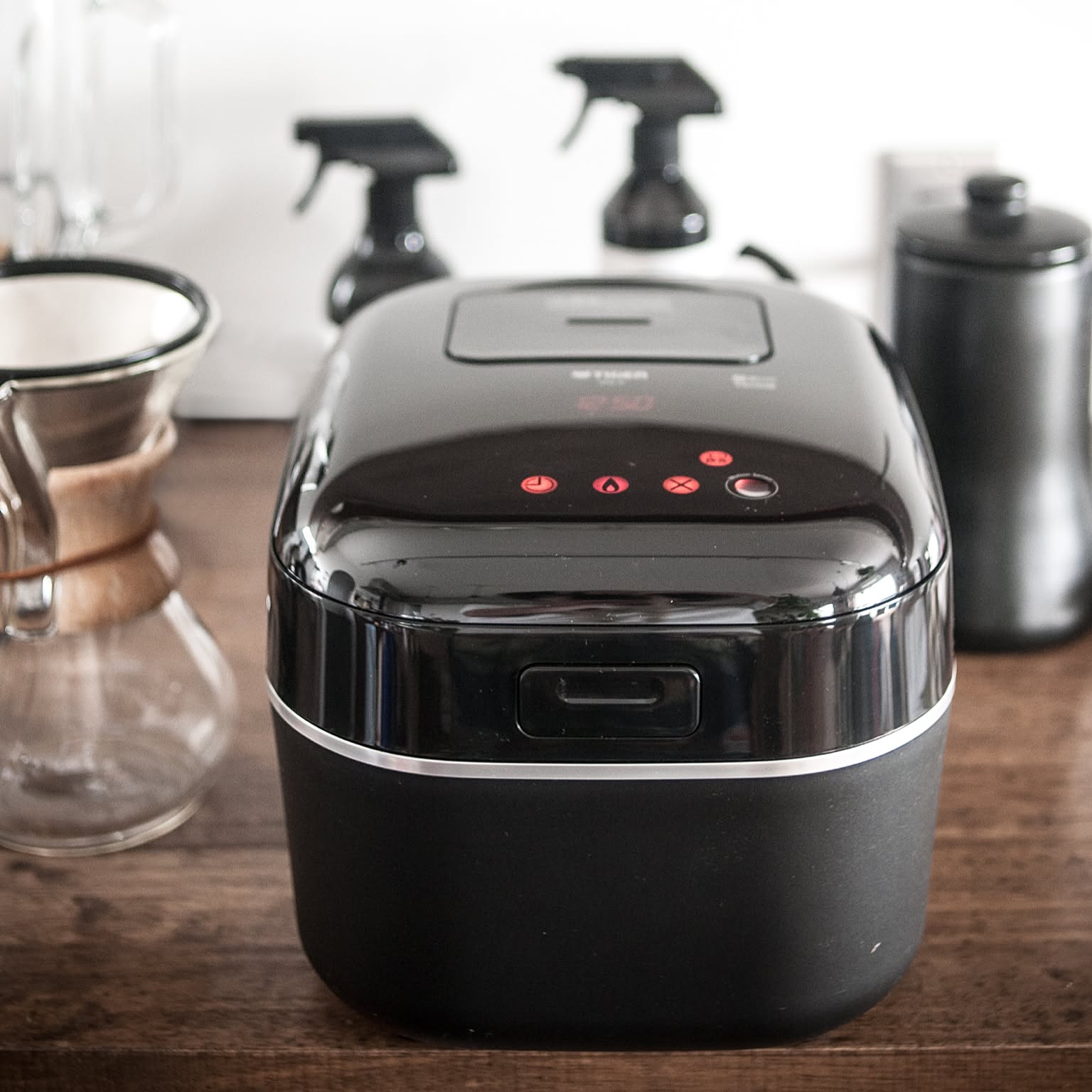 A glossy black Zojirushi rice cooker shown front and center on a dark wooden counter beside coffee gear.