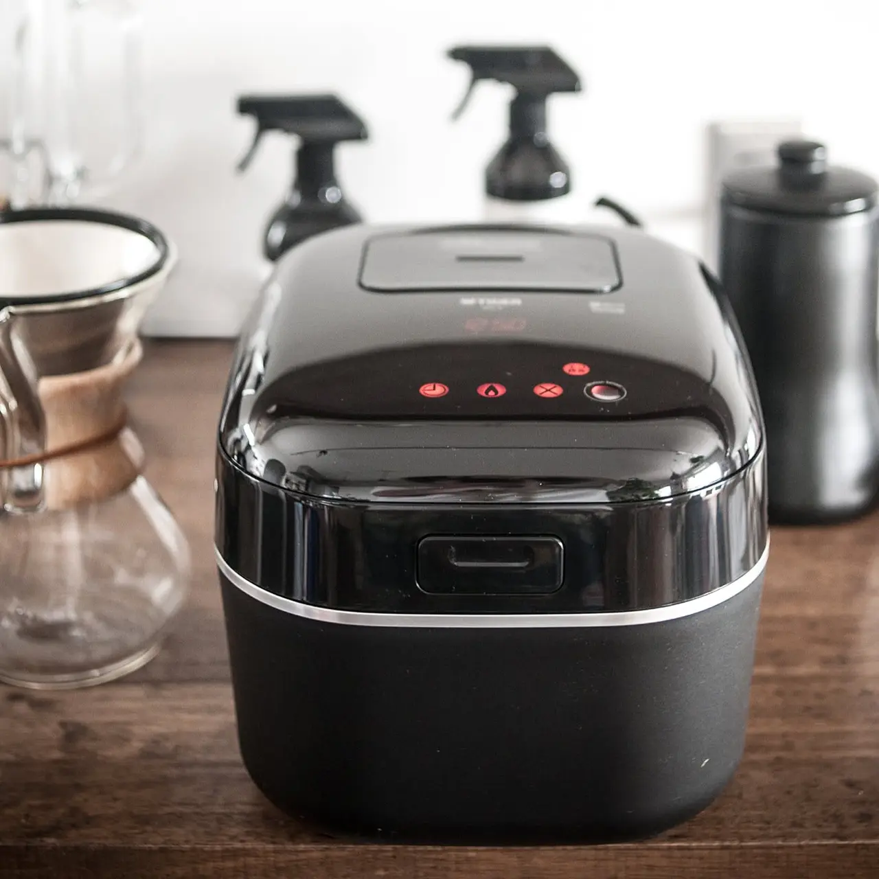 A glossy black Zojirushi rice cooker shown front and center on a dark wooden counter beside coffee gear.