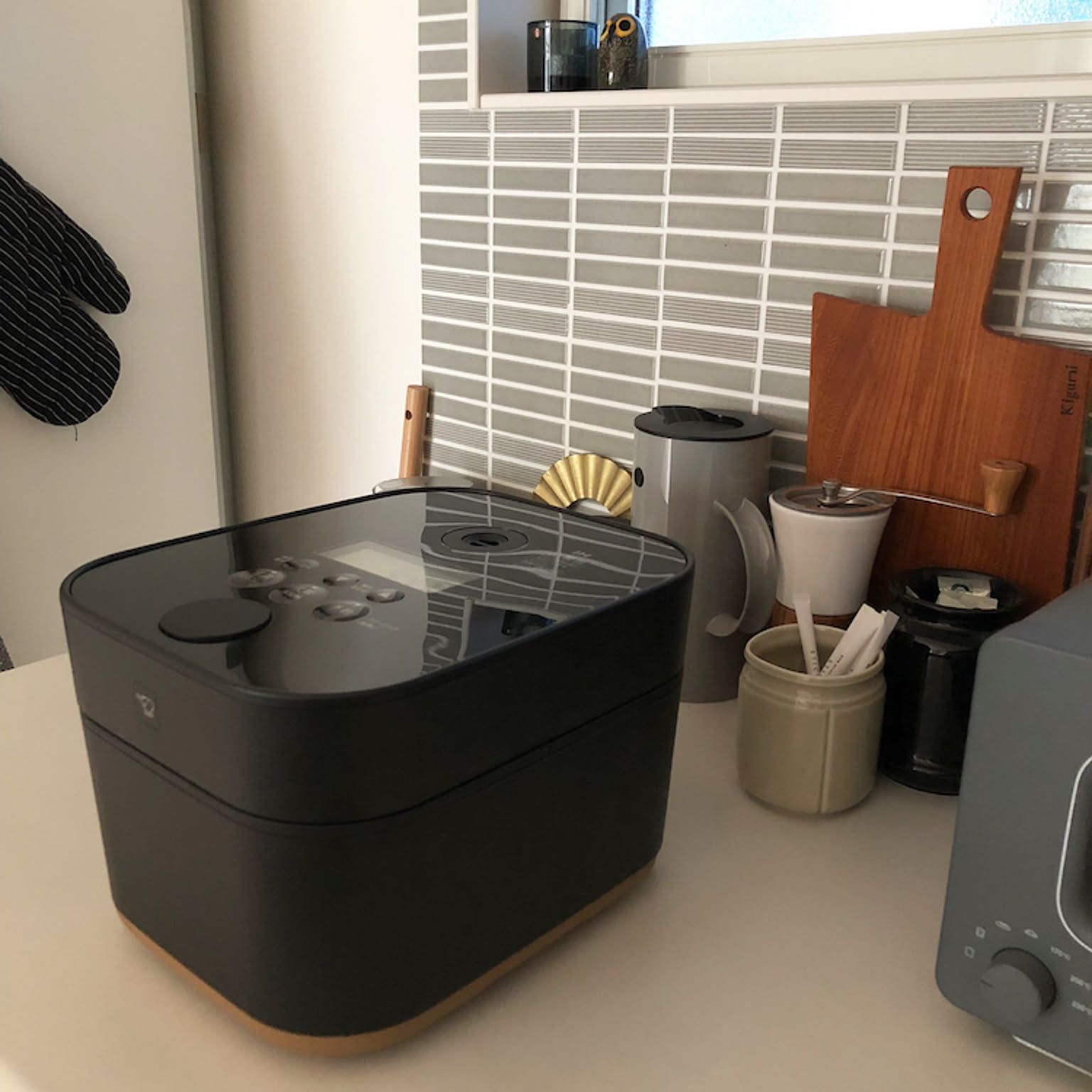 A Zojirushi STAN. rice cooker in matte black sitting on a kitchen counter alongside cutting boards and kitchen tools in a modern Japanese home