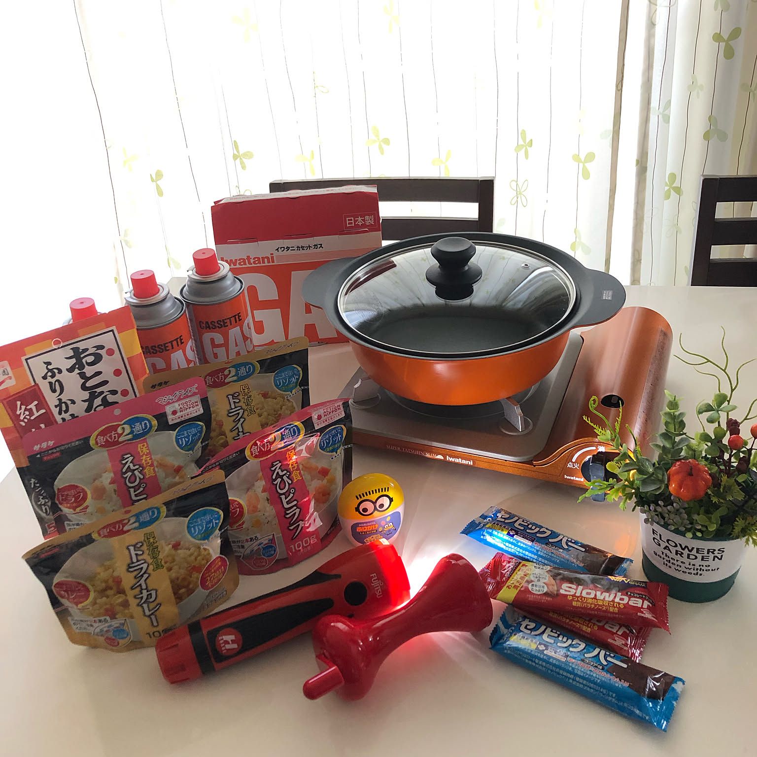 A compact Iwatani cassette stove displayed with a pot, fuel canisters, and shelf-stable food for an indoor emergency cooking setup.