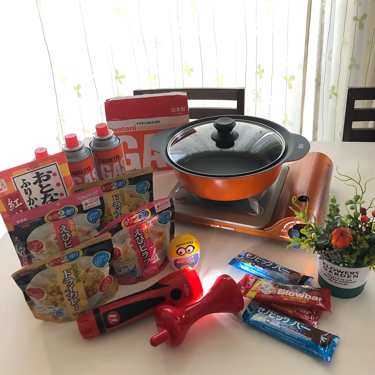 A compact Iwatani cassette stove displayed with a pot, fuel canisters, and shelf-stable food for an indoor emergency cooking setup.