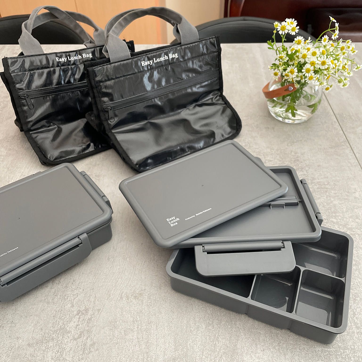 Several gray adult bento boxes with removable dividers and black insulated lunch bags on a table.